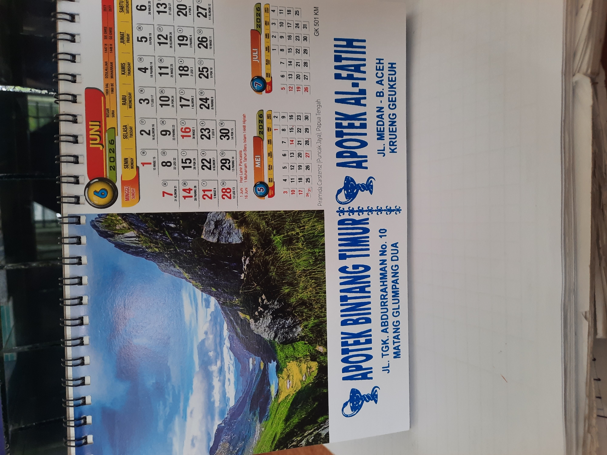 The image shows a calendar with the month of June 2026 and July 2026. The calendar is open, revealing the monthly layouts. Below the calendar, there is text indicating two pharmacy names: "APOTEK BINTANG TIMUR" and "APOTEK AL-FATIH".
The address for "APOTEK BINTANG TIMUR" is "JL. TGK. ABDURRAHMAN No. 10 MATANG GLUMPANG DUA".
The address for "APOTEK AL-FATIH" is "JL. MEDAN - B. ACEH KRUENG GEUKEUH".
Matang Glumpang Dua is a village in the Lhokseumawe city of Aceh, Indonesia. Krueng Geukueh is also a subdistrict in the Lhokseumawe city, Aceh, Indonesia. The presence of these two addresses on a single calendar suggests that either these are two branches of the same pharmacy or they are collaborating in some way. The calendar also features a scenic mountainous landscape with a lake and some vegetation.