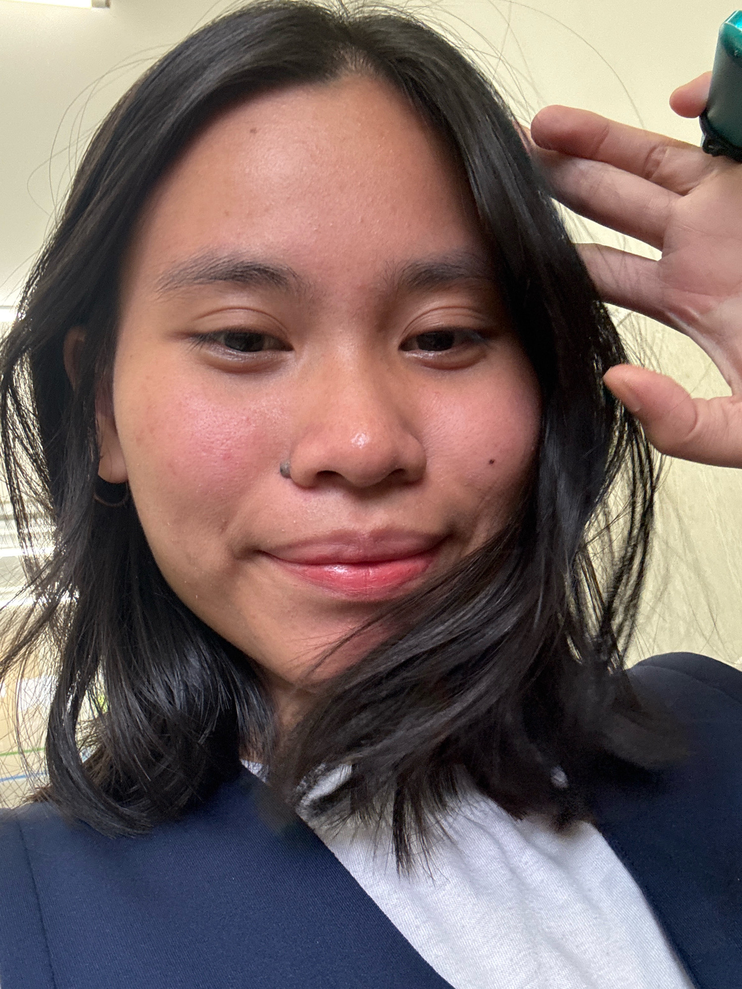 A young woman with shoulder-length, dark hair and a fair complexion is shown in a close-up, selfie-style shot. She is wearing a dark navy blazer over a white collared shirt. Her right hand is raised, with fingers spread, near her face. A dark, small object is visible on her right cheek near her nose. Her lips are glossy and slightly parted, and she appears to be smiling faintly. The background is a light-colored, textured wall. The image does not provide any specific details indicating its location in East Jakarta, Indonesia, or any urban setting.