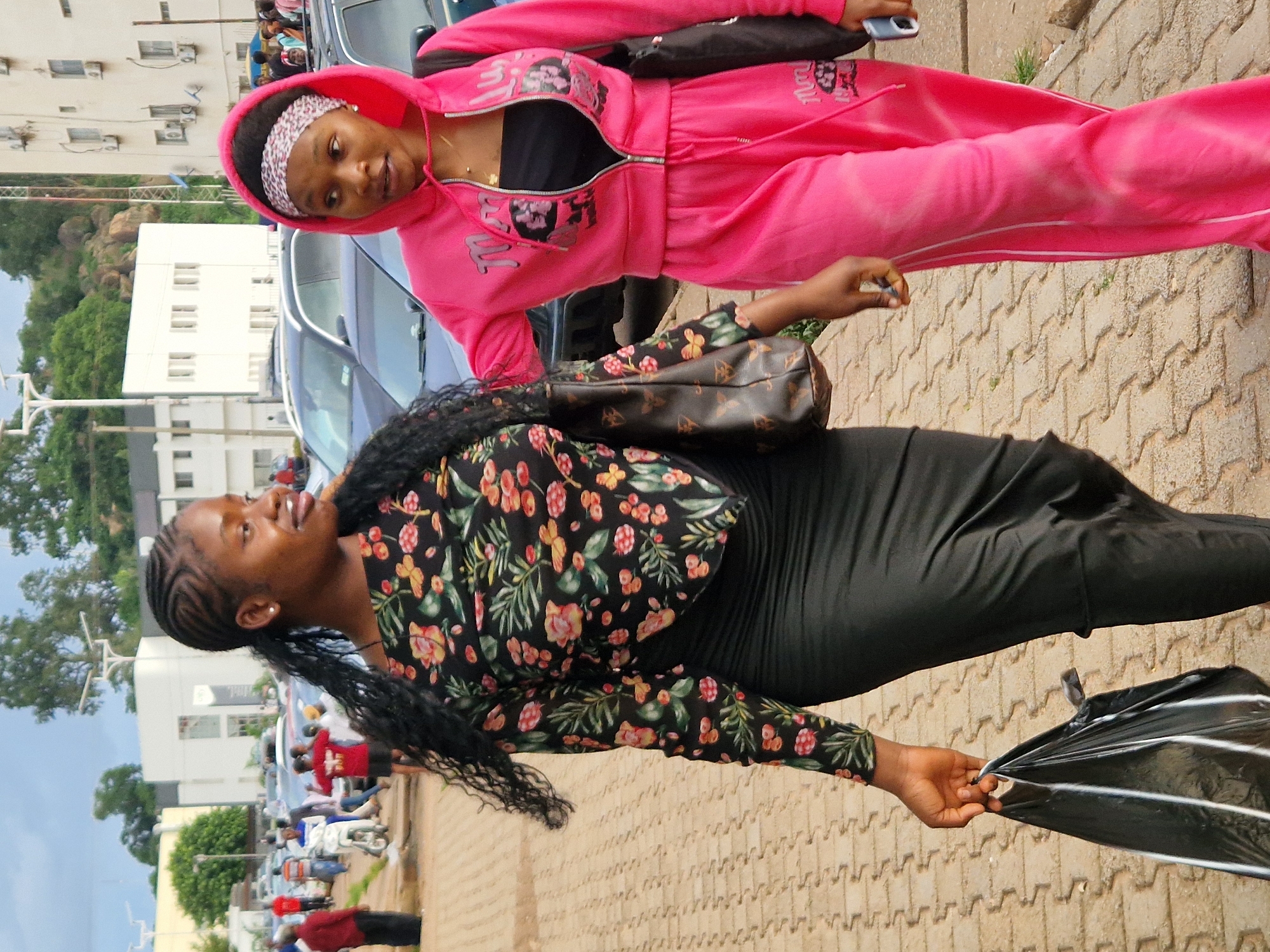 Two young women are standing outdoors on a paved walkway. The woman in the foreground is wearing a black long-sleeved dress with a floral pattern and a black skirt. She has braided hair that falls down her back and is holding a black umbrella in her right hand. She is looking up and to her right with a slight smile. The woman in the background is wearing a bright pink tracksuit and a black top underneath. She has a white and pink patterned headband on. She is looking forward with a neutral expression. Behind the women, there are cars and buildings, suggesting an urban environment. The time of day appears to be daytime, and the weather seems pleasant. The setting is likely a public space in Jos, Nigeria, given the context provided. There is no visible text in the image.