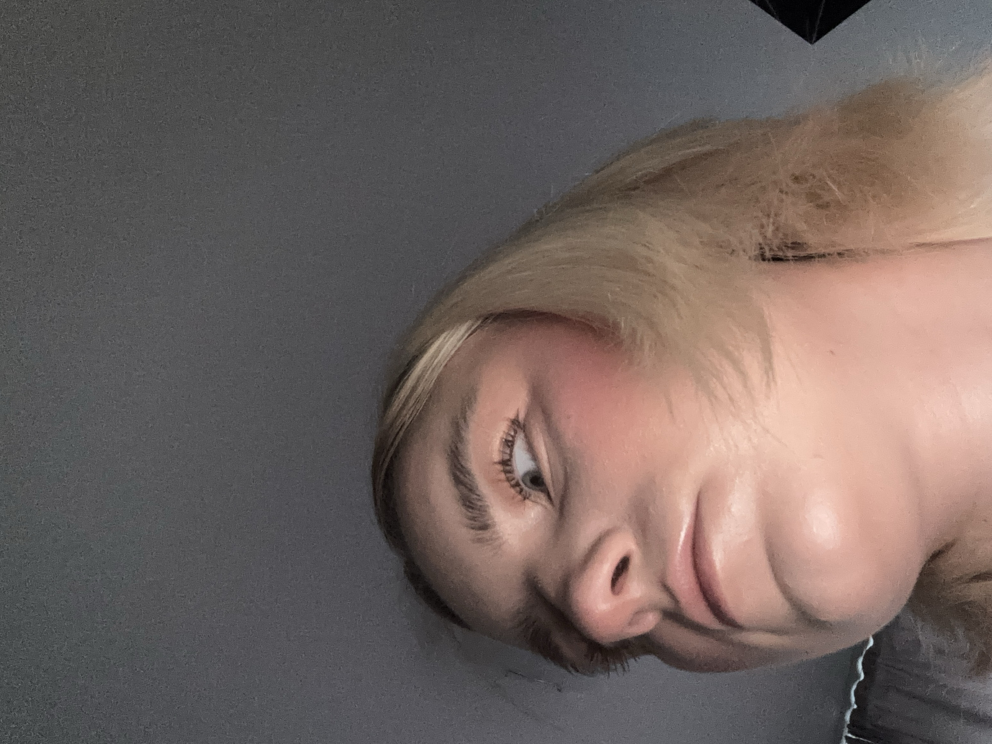 A close-up, angled selfie of a fair-skinned individual with blonde hair. The person's face is prominently featured, showing one eye, nose, and mouth. Blush is visible on the cheek. The background is a plain, dark gray wall. No information indicates this image was taken in Vsevolozhsk, Russia.