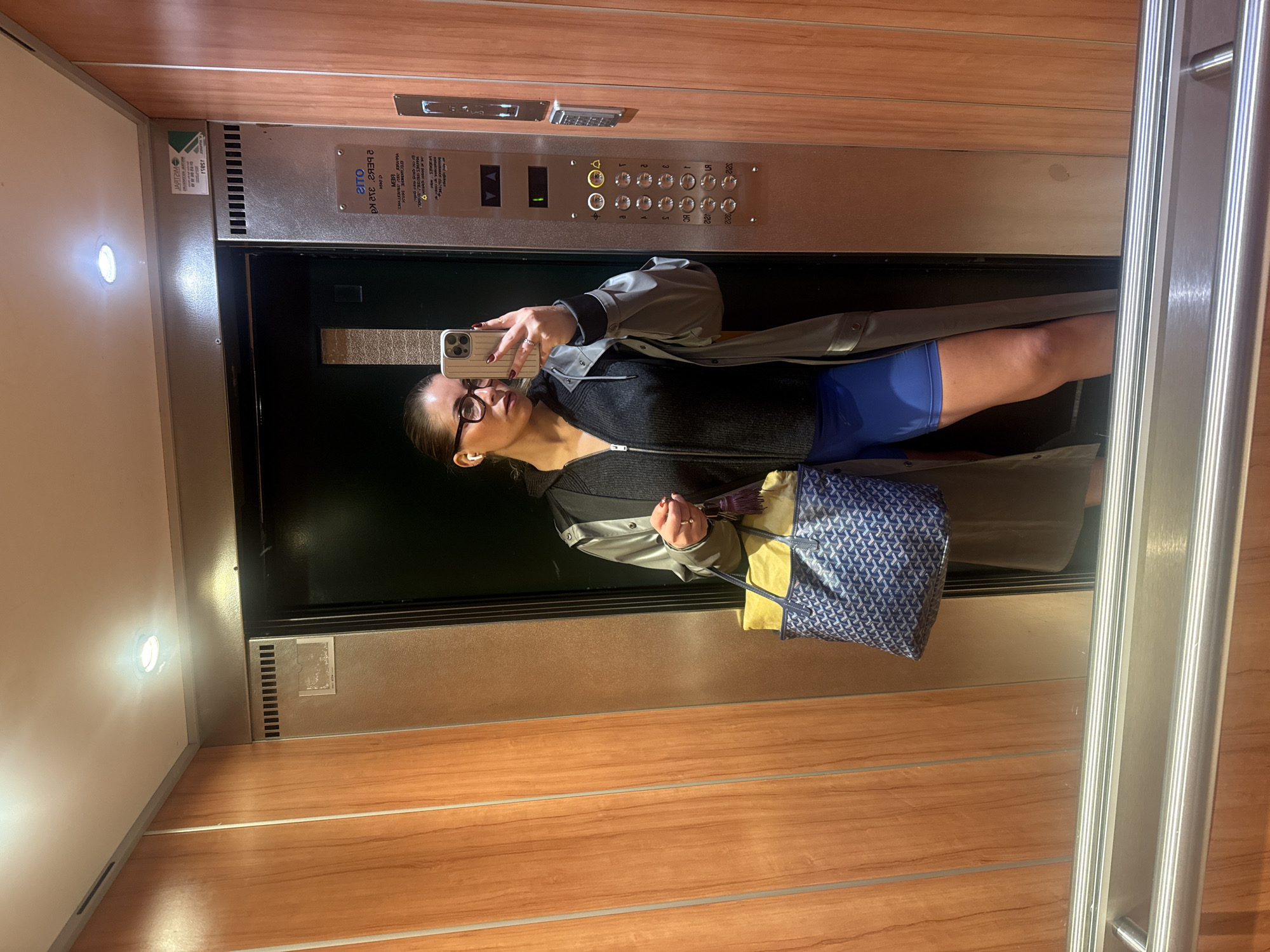 A woman is taking a selfie in an elevator. She is wearing glasses, a black sweater, blue shorts, and a grey trench coat. She is holding a blue patterned tote bag with yellow accents. The elevator has wood-paneled walls and a metal control panel with floor buttons. The city is not determinable from the image.