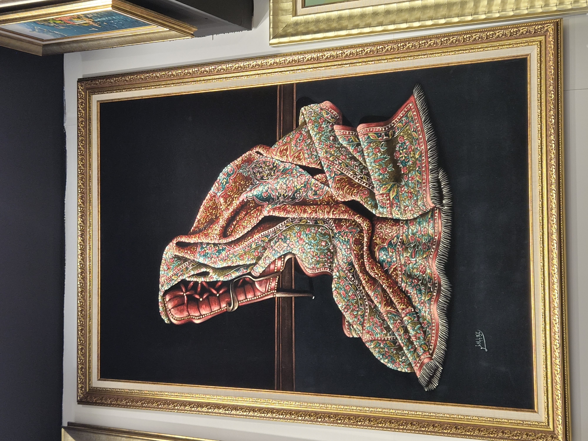 The image shows a framed painting of a richly patterned rug draped over a red upholstered chair against a dark background. The rug features intricate floral designs in vibrant colors such as red, pink, green, and yellow, with a fringed edge. The chair appears to be made of leather or a similar material, with tufted detailing. A dark wooden shelf or ledge is visible behind the chair. The painting is signed in the lower left corner with Arabic script. The frame is ornate and gilded. The painting is displayed in an indoor setting, likely an art gallery or shop, with other framed artwork visible to the right. The location is identified as Maltepe, Turkey.