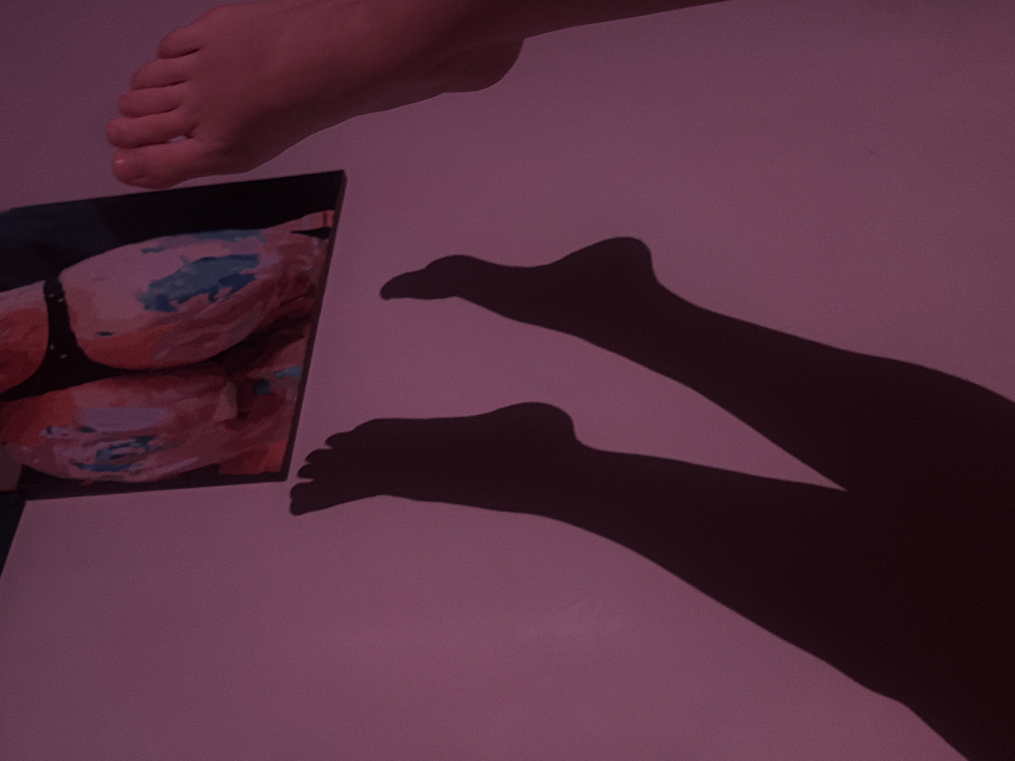 The image shows a pair of feet in the upper part of the frame, close to a framed picture. The framed picture has an abstract design featuring orange and blue colors. Below the feet, a shadow of legs is cast onto a wall or surface, lit with a pinkish light. The location is not discernible from the provided image.