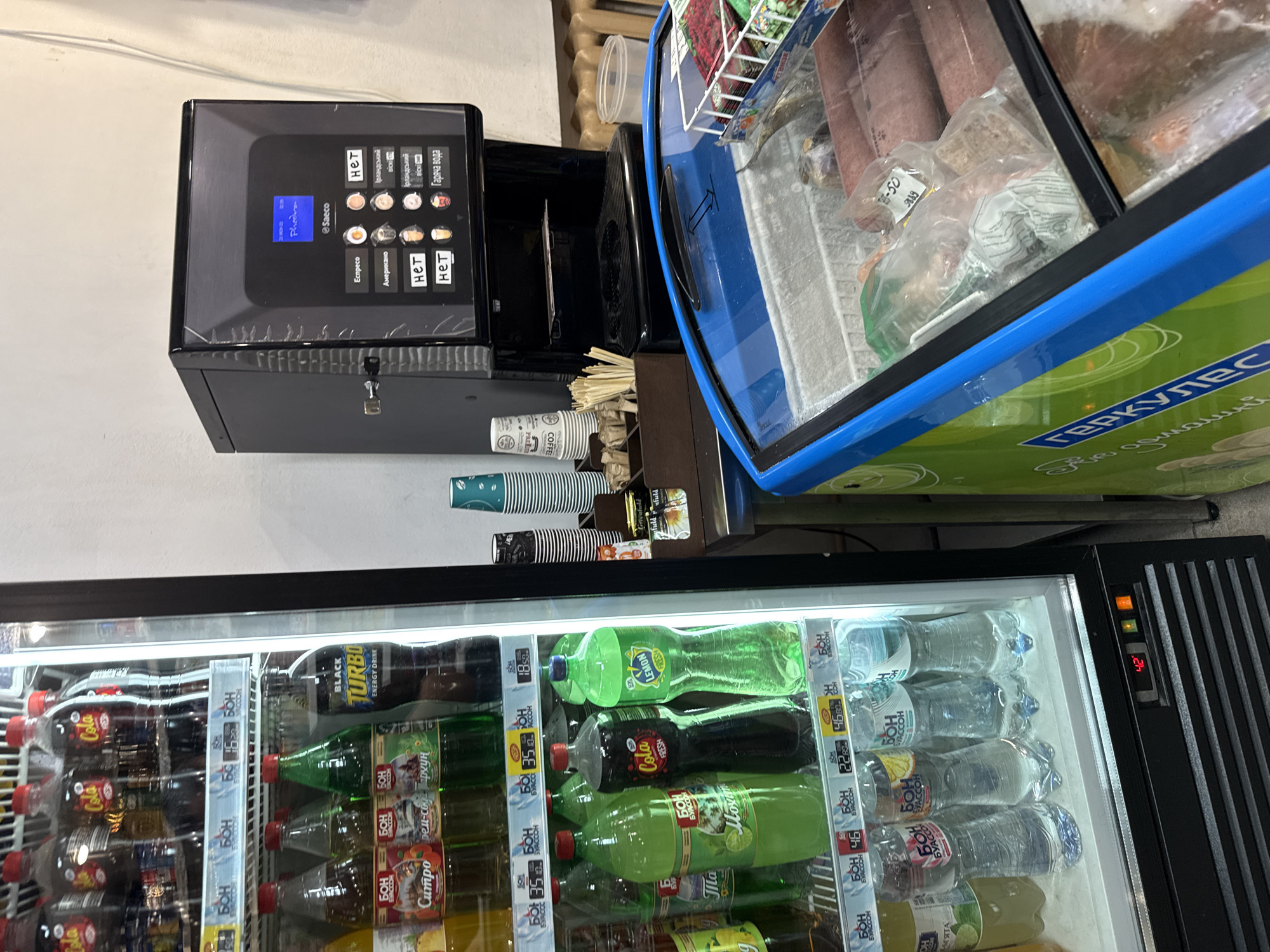 The image captures an indoor commercial setting in Donets, Ukraine, likely a small store or self-service area. The scene features a coffee vending machine, an open-top freezer, and a large upright glass-door refrigerator filled with beverages.
In the upper left, a black Saeco Phedra coffee machine displays selection options on its screen. Visible text includes "Ecпpeco" (Espresso), "Американо" (Americano), "Гаряча вода" (Hot water), and "Ірландський" (Irish), with some options marked "нет" (not available). Beside the machine are stacks of disposable cups, some bearing "COFFEE" and "Greenfield" branding, along with stirrers.
To the right of the coffee machine, an open-top freezer holds various plastic-wrapped frozen food items. A price tag with "-50" and "9889" is visible on one package. The side of this freezer features the text "ГЕРКУЛЕС" (Hercules).
Dominating the lower half of the image is a brightly lit glass-door refrigerator packed with bottled drinks. Various brands and types are discernible, including "Cola", "BLACK TURBO ENERGY DRINK", "LEMON FRESH", "Cola FRESH", "МЯТА" (Mint), and "Ситро" (Citro). The brand "БОН КВАССОН" (BON KVASSON) appears frequently on price tags, which show values like "18.50", "35", "46", and "22.50". The refrigerator's control panel displays a temperature of "4.2". No people are present, depicting a quiet retail setup.