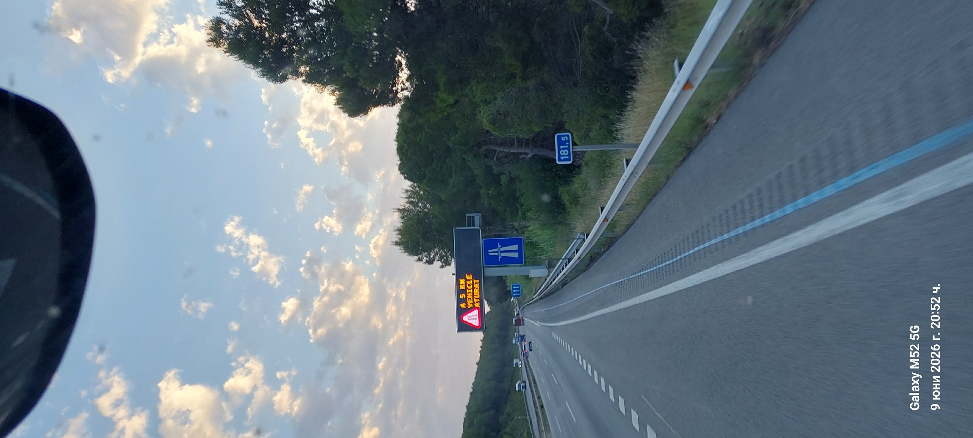 The image shows a highway scene in Subirats, Spain, captured from a vehicle's perspective. The sky is partly cloudy with warm, golden light suggesting it's either sunrise or sunset. A digital traffic sign is prominently displayed, showing a red triangular warning symbol and the text "A 5 KM VEHICLE ATURAT". Below this sign is a blue highway directional sign with a white depiction of a bridge or overpass. To the right, a blue circular sign indicates "181.5". The highway has multiple lanes marked by white lines, and a silver guardrail runs along the right side of the road. The surrounding environment is characterized by lush green trees and vegetation lining the highway. There are several vehicles visible on the road ahead. The bottom left corner of the image contains a timestamp and device information: "Galaxy M52 5G 9 юни 2026 г. 20:52 ч.".