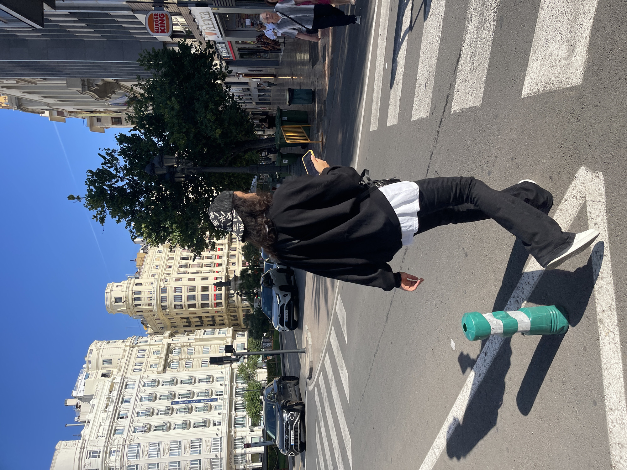 The image captures a sunny urban scene in València, Spain, specifically near the "Hotel Venecia" and "Plaza Centro" as indicated by banners on a building in the background. A person with long, dark, curly hair and a black New York Yankees baseball cap walks across a zebra pedestrian crossing from left to right, facing away from the camera. They are dressed in a black long-sleeved top layered over a white shirt, black pants, and white shoes, while looking at a yellow smartphone held in their right hand. A black bag is visible on their left hip.
In the immediate foreground, a green and white striped flexible traffic bollard stands on the asphalt road, casting a long shadow to the left. The pedestrian crossing stripes are prominent across the width of the frame. In the mid-ground on the left, a dark grey Volkswagen T-Roc SUV (license plate 7088 WKJ) is positioned at an intersection. Behind it, a white Toyota RAV4 SUV (license plate 3632 NJC) is also visible. A traffic light pole stands near the center of the intersection.
The background features multi-story, light-colored buildings with ornate architecture. On the left, a large building with numerous balconies and intricate details is identified as "Hotel Venecia" and "Venecia Plaza Centro" by blue banners. Further back on the left, other classical buildings with rounded turrets are visible under a clear blue sky. On the right side of the street, several retail establishments are present, including a "Burger King" with its distinctive orange sign and an "I ❤️ Valencia" sign below it. Further down this street, an older man in a light-colored shirt and dark pants, carrying a shoulder bag, walks towards the camera on the sidewalk, accompanied by another person partially obscured. Green waste bins and yellow barriers are also visible along the sidewalk on the right. Trees with green foliage are scattered throughout the scene, and strong sunlight casts distinct shadows.