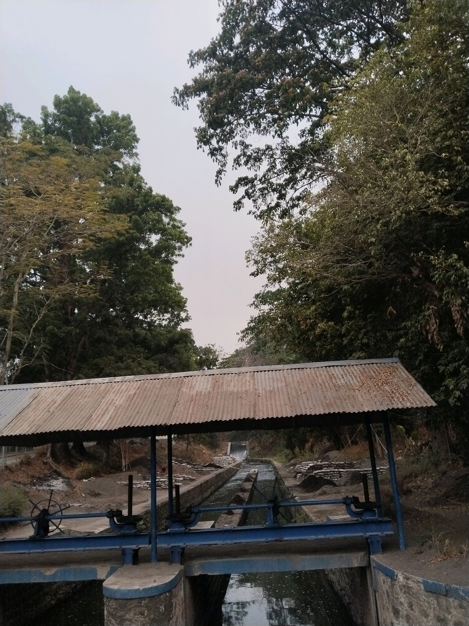 The image depicts an outdoor scene in Sangkanjaya, Indonesia, featuring a canal system with sluice gates. A weathered, corrugated metal roof supported by blue metal posts covers a portion of the canal and its control mechanism. The sluice gate mechanism, painted blue, includes hand wheels used for operation. Water flows through the concrete and stone canal, which is flanked by dirt pathways and vegetation. Large trees with dense foliage dominate the background, creating a shaded environment. The sky appears overcast. There are no people visible in the frame.