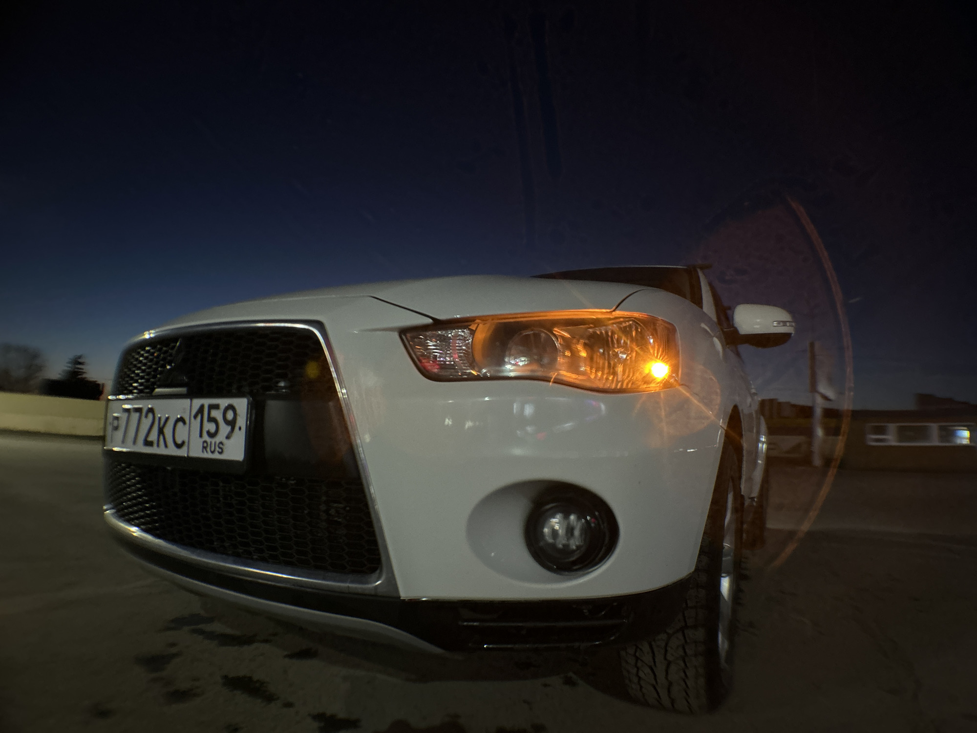 A low-angle, fisheye-lens photograph taken at night features a white Mitsubishi Outlander SUV. The vehicle displays the license plate P772KC 159 RUS, indicating Russian registration. The right headlight and fog light are illuminated. The vehicle is parked on a paved surface in the city of Chaikovsky, Russia. A low building with illuminated windows is visible in the background. The sky ranges from a deep blue to near black.