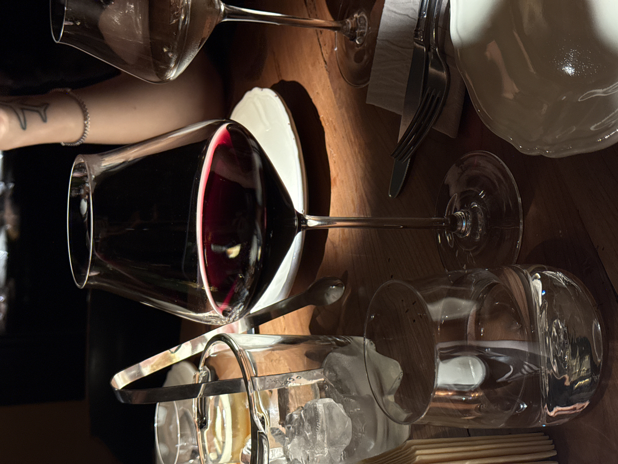 The image depicts a restaurant table setting, potentially in Moscow, Russia, though that cannot be confirmed from the image alone. The table, made of wood, is set with wine glasses, one filled with red wine, another empty. A white plate rests near the red wine glass. Silverware (fork and knife) lies on a napkin. A container of ice with tongs is also present. An arm with a tattoo and bracelet is partially visible. The overall impression is of a dining scene.