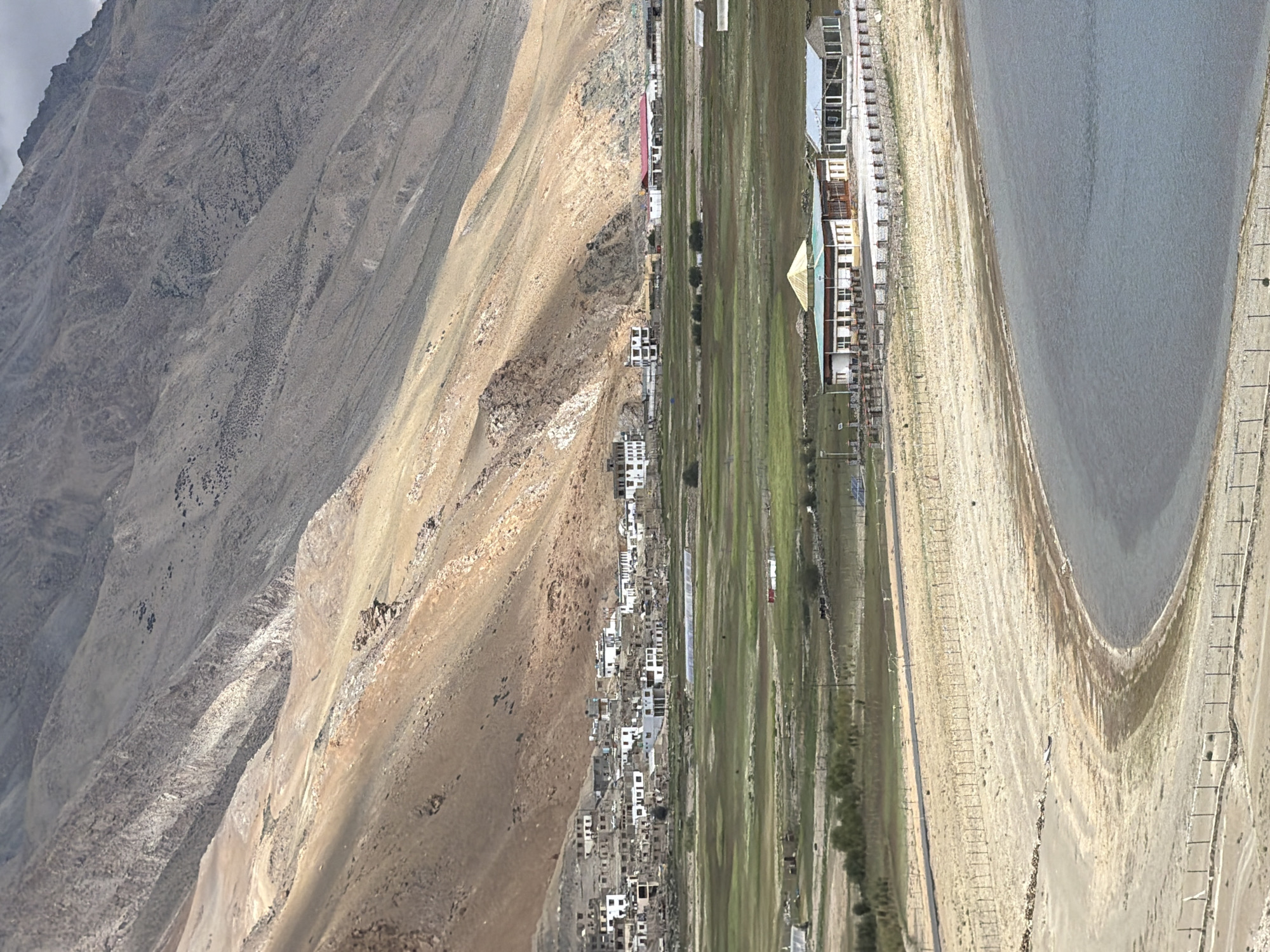 The image shows a panoramic view of the town of Karzok, India, situated in a valley at the base of a large, arid mountain range. The mountain slopes are characterized by varying shades of brown and grey, with sparse vegetation visible on the lower sections. In the middle ground, a cluster of white and beige buildings forms the village of Karzok. These structures appear to be residential or communal buildings, with some featuring flat roofs and multiple stories.
In front of the village, a vast expanse of green fields stretches out, dotted with a few scattered buildings and what appear to be greenhouses. A road runs through this agricultural area, connecting to the village. Closer to the foreground, a curved body of water, likely a lake or reservoir, occupies the lower right portion of the image. The shoreline is a sandy, dry expanse bordered by a fence. Further into the foreground, another fenced area encloses a building complex with a prominent green-roofed structure and adjacent glass-enclosed buildings. The sky is overcast with scattered clouds. The overall scene depicts a remote, high-altitude settlement surrounded by natural landscapes.