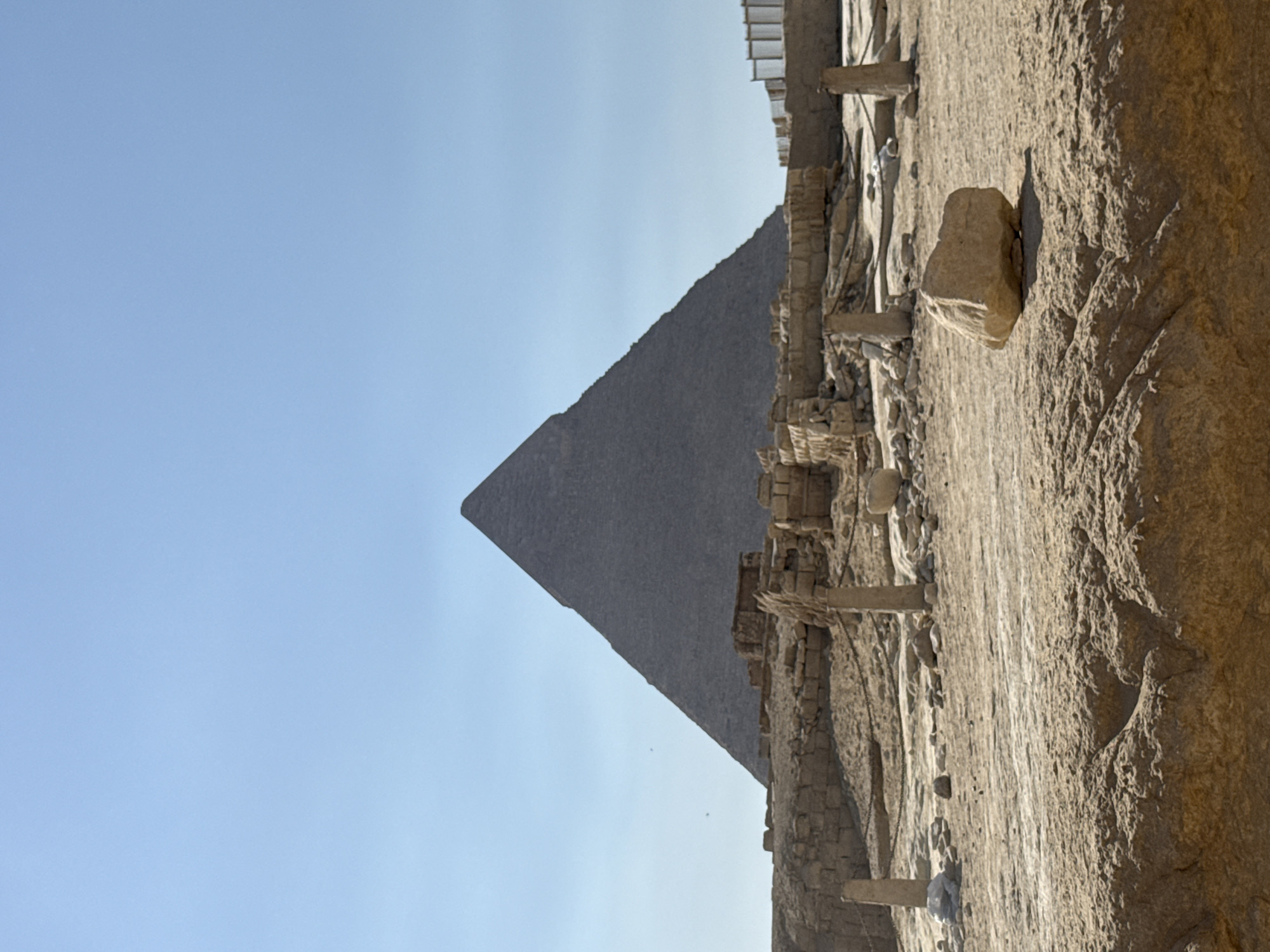 The image depicts the Great Pyramid of Giza, a monumental ancient structure located in Giza, Egypt, on the outskirts of Cairo. The pyramid, made of rough-hewn stone, dominates the background under a clear, pale blue sky. In the foreground, the sandy terrain is scattered with rubble, remnants of ancient stone walls, and a single large, light-colored rock. The area appears to be an archaeological site, with further stone structures and barriers visible to the right. There are no people or animals visible in the scene. The lighting suggests a bright, sunny day.