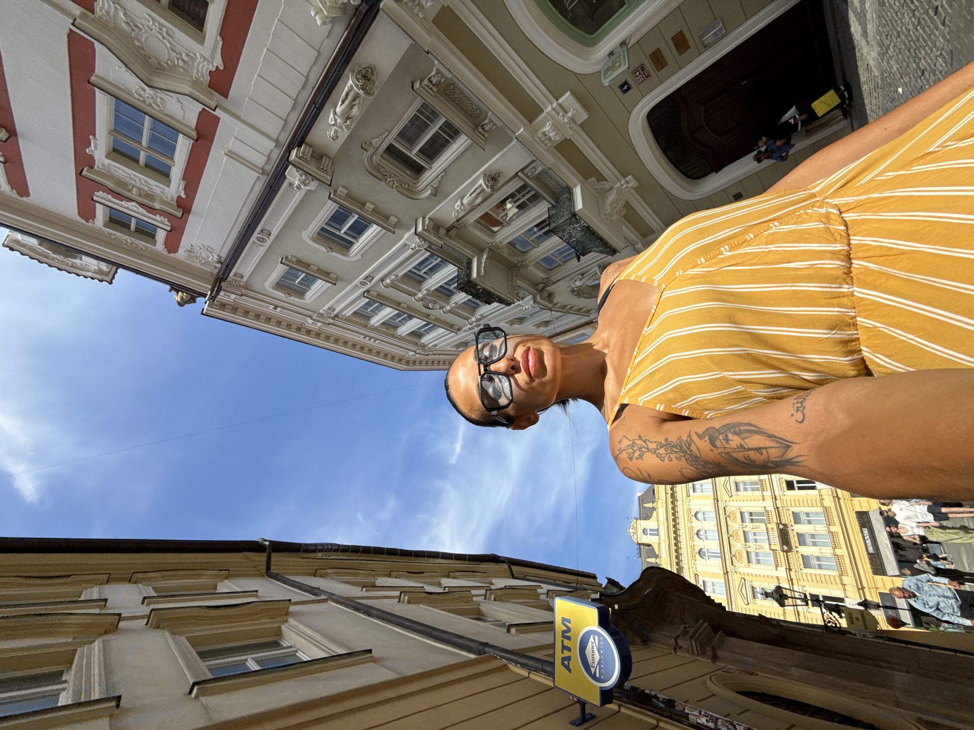 A first-person perspective low-angle selfie shows a woman with dark hair and sunglasses standing on a cobblestone street in Prague, Czechia. She is wearing a yellow and white striped dress and has a tattoo on her left arm. Behind her, on the left side of the frame, is a yellow ATM sign. The street is lined with buildings. The building on the right has intricate architectural details and is painted in shades of cream and red. The building on the left appears to be under renovation, with scaffolding visible. The sky is blue with scattered clouds. In the background, several people are walking on the street.