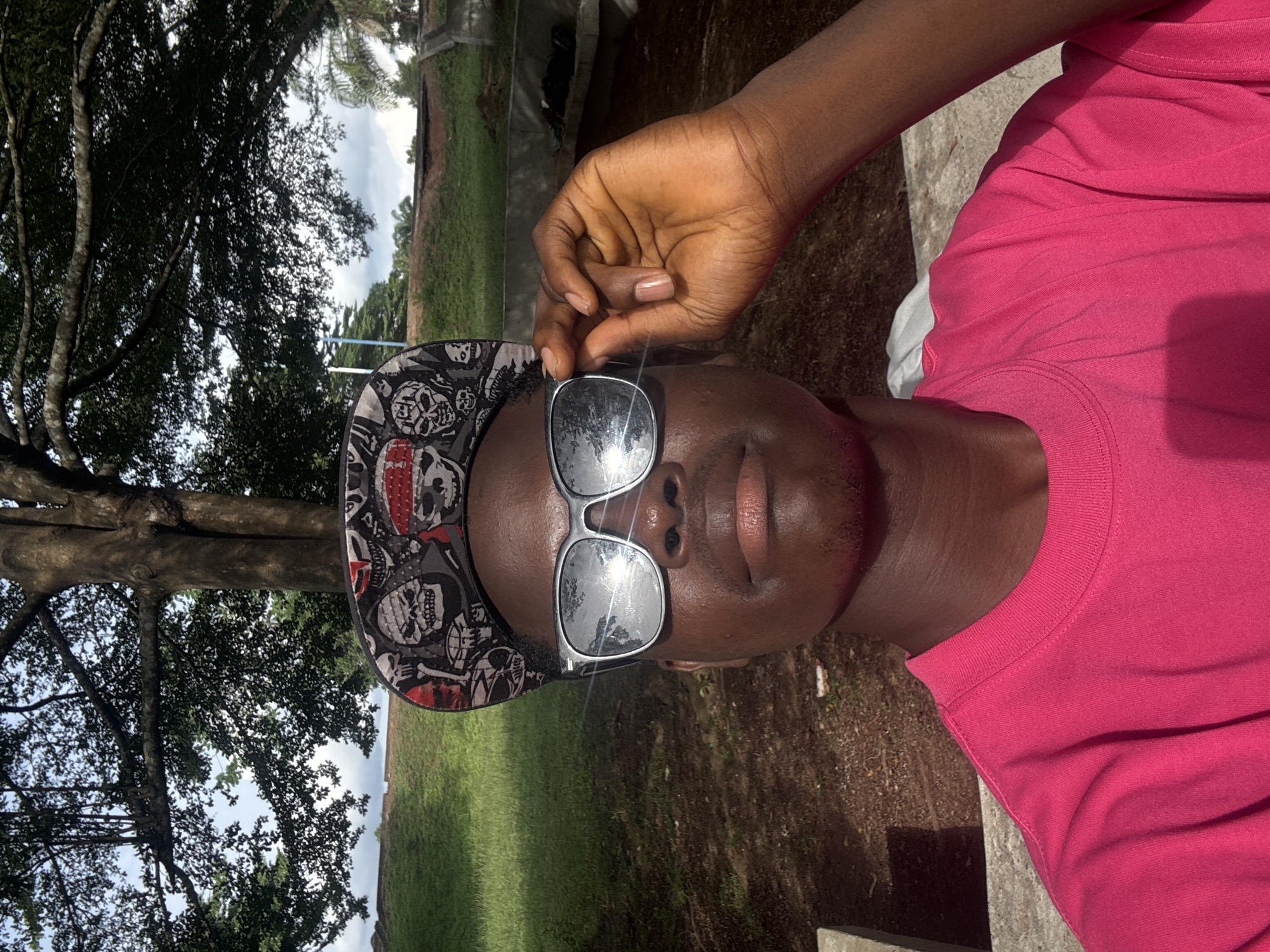 A medium shot shows a dark-skinned person wearing a pink T-shirt, a skull-printed hat, and sunglasses in Ihiagwa, Nigeria. The person's right hand is near their sunglasses. A tree, green grass, and a cloudy sky are visible in the background. The photo appears to be taken outdoors on a partially sunny day.