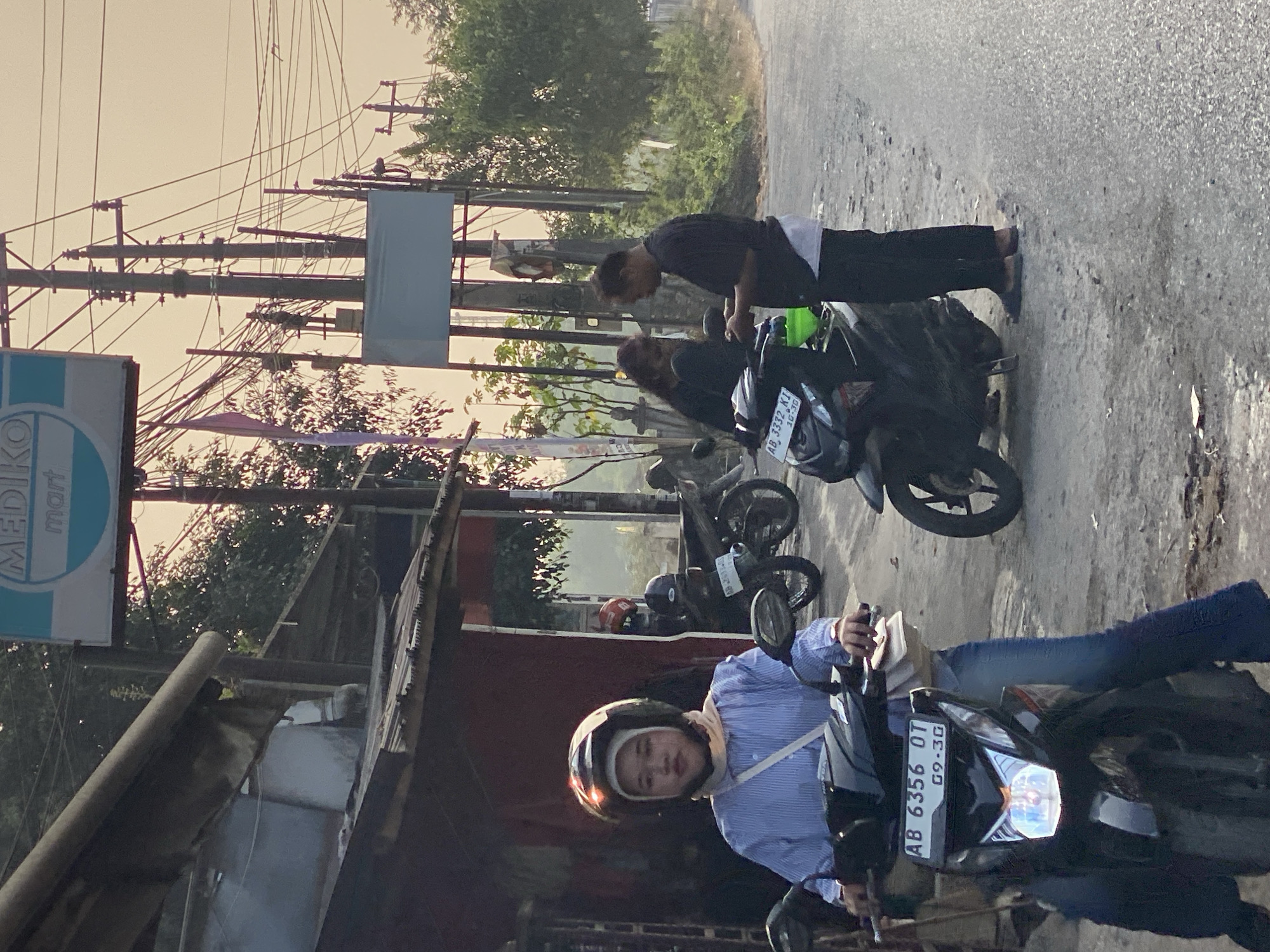 In Kasihan, Indonesia, a woman wearing a blue and white striped shirt and a hijab sits on a black scooter. She has a light beige scarf around her neck and a white strap across her chest. The scooter's license plate reads "AB 6356 OT G9-30". Behind her, a man in a black t-shirt and white shorts stands next to another black scooter, interacting with a woman who is partially obscured. The background features a street with power lines and poles, lush green trees, and a sign for "MEDIKO mart". The scene appears to be taking place in the late afternoon or early evening.