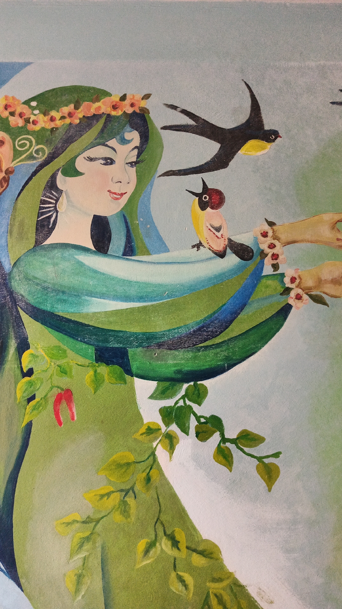 The image is a painting featuring a woman with dark hair adorned with a floral crown and a crescent-shaped ornament on her forehead. She is wearing flowing green and blue garments. Two birds are depicted in the scene: a larger black and yellow bird in flight and a smaller bird perched on the woman's outstretched arm. The woman's arms are extended, and two hands, possibly also belonging to the woman or another figure, are visible on the right side, decorated with small pink flowers. Green leaves and red peppers are also incorporated into the composition, suggesting a natural or harvest theme. The background is a soft gradient of light blue and green. The painting does not depict any recognizable landmarks or features that would allow for its specific location within the city of Kryvyi Rih, Ukraine.