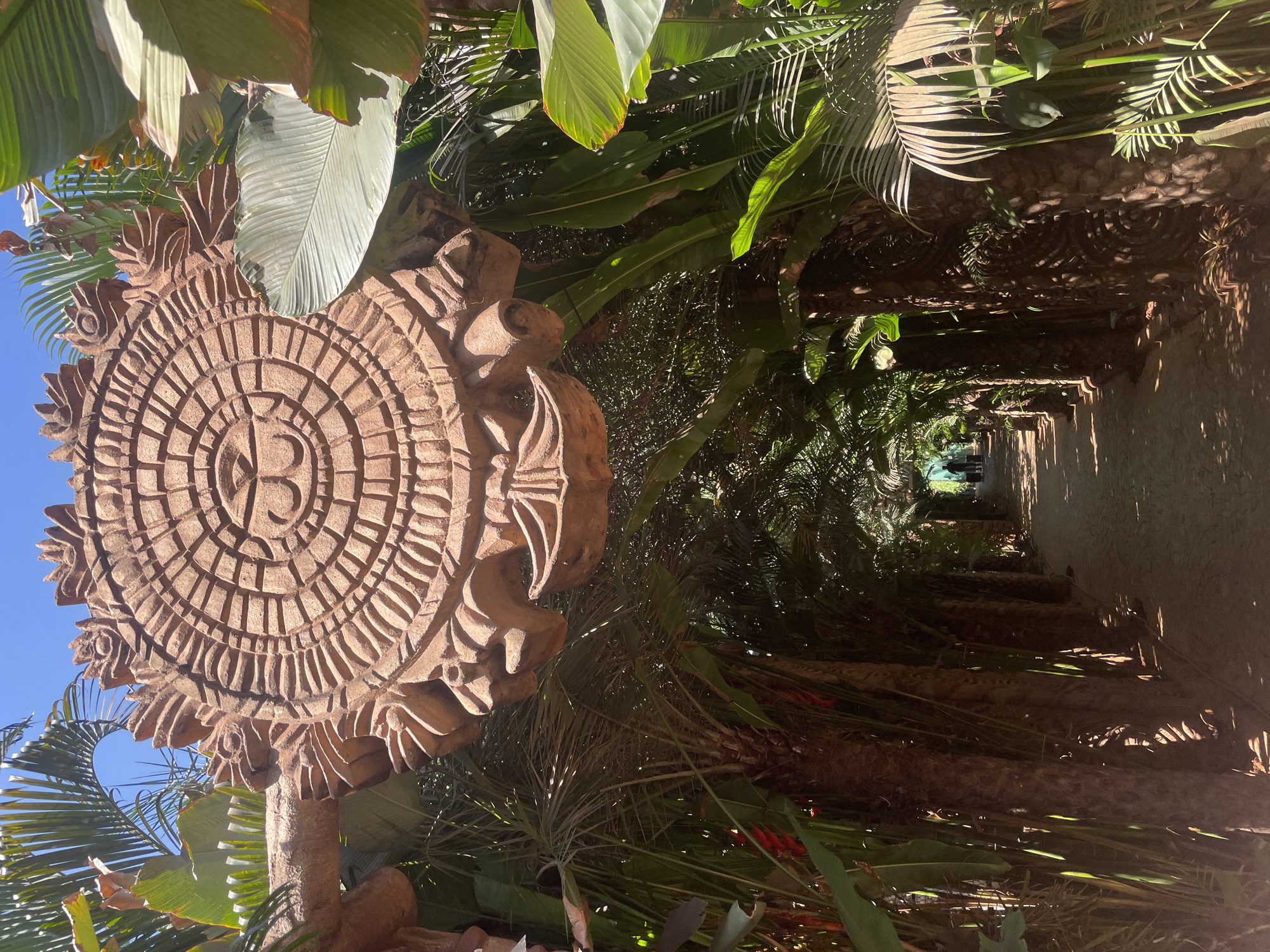 The photograph depicts a pathway in Ashvem, India. The pathway is bordered by lush tropical foliage and leads towards a brighter, indistinct area in the distance where the silhouettes of two people can be seen. A large, intricately carved stone sculpture dominates the left foreground. The sculpture is round and features concentric circles and geometric patterns. The sculpture is also partially obscured by leaves and palm fronds, suggesting a well-established, natural environment. The lighting creates strong contrasts and shadows along the pathway and sculpture.