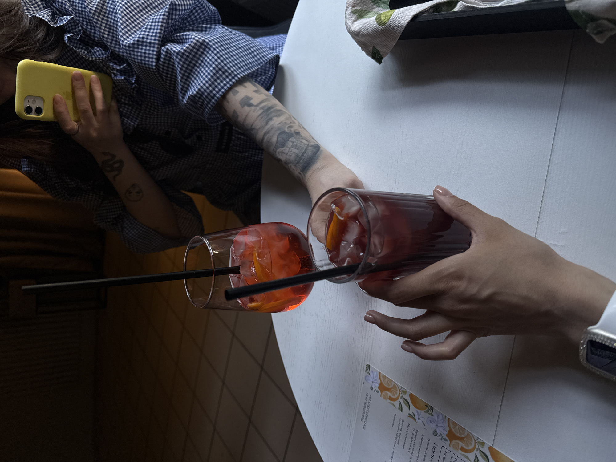 The image depicts a social scene, possibly at a restaurant or bar. Two individuals are present at a white, round table. One person is holding a yellow smartphone, wearing a blue and white checked shirt, and has tattoos on their arm. Another person, wearing a white shirt and a smartwatch, is holding a glass. Two glasses with orange-colored drinks and black straws are on the table. A menu with Russian text is also visible on the table. The OCR text in the image indicates this may be a cafe in Votkinsk, Russia.