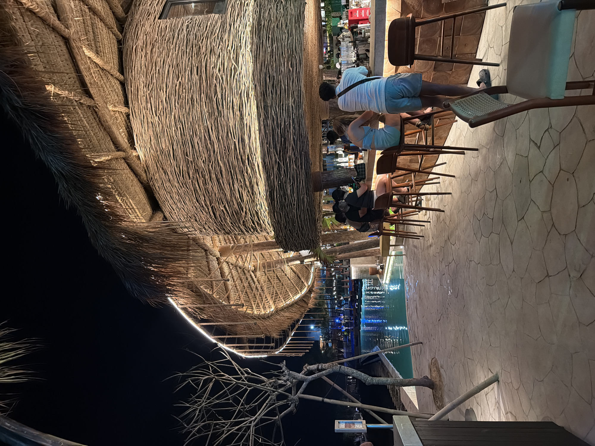 This image depicts an outdoor bar or restaurant area at night, likely in North Kuta, Indonesia, given the location context. The central feature is a large, unique structure on the left, resembling a massive bird's nest or a traditional hut, constructed from intricately woven dried reeds, branches, or thatch. This structure is warmly illuminated by ambient lighting and a distinctive white LED strip tracing its curved outline.
Several people are visible, seated on high wooden stools at a long bar counter positioned to the right of the main structure. They appear to be casually dressed, socializing, and enjoying drinks. Behind the bar, bottles and glassware are arranged, and more patrons can be seen in the background.
Adjacent to the bar area and running along the right side of the scene is a body of water, likely a swimming pool or decorative pond, which reflects blue lights from beneath its surface. A paved pathway with an irregular stone pattern borders the water and the bar. In the foreground on the left, the bare, intertwining branches of a tree stand out against the dark night sky. The overall lighting scheme is warm and inviting, highlighting the natural textures of the construction materials. The time of day is clearly night, with the darkness emphasizing the artificial illumination. A small, indistinct sign is visible in the lower right foreground, but its text is illegible. The setting conveys a relaxed, tropical, and leisurely atmosphere.