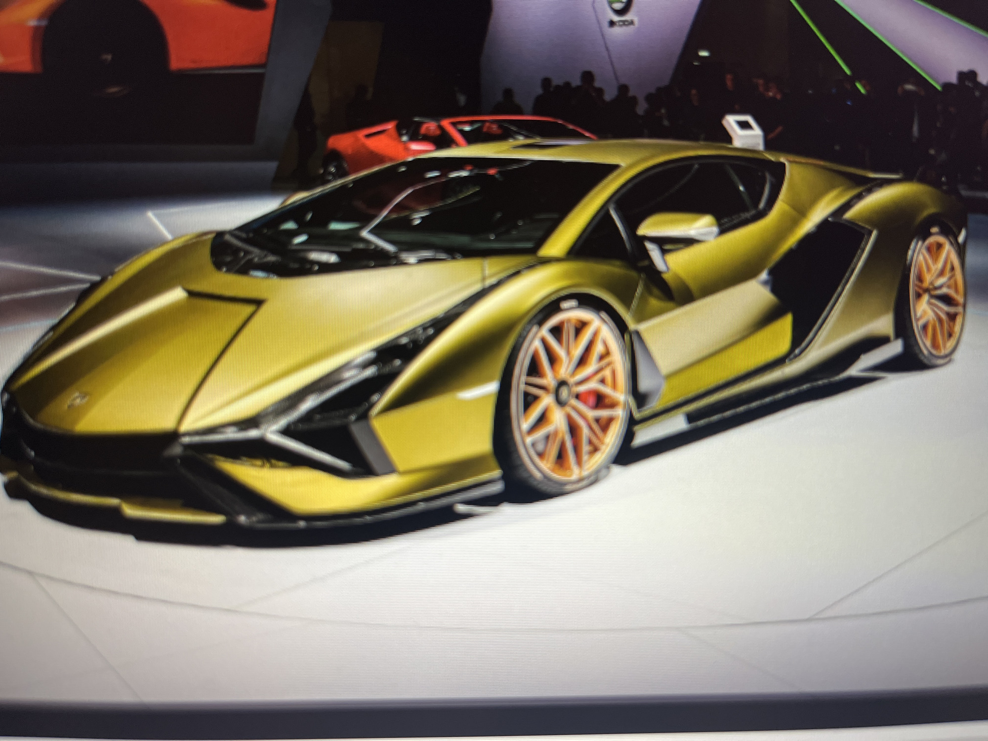 The image displays a striking golden supercar, likely a Lamborghini Sian, as the main subject at an indoor car show or automotive exhibition. The car is centrally positioned on a light-colored platform, its futuristic and angular design highlighted by focused lighting. Its distinctive features include sleek lines, aggressive front fascia, and unique orange/bronze multi-spoke wheels with visible red brake calipers.
In the background, two red sports cars are partially visible, one prominently behind the golden car and another further back to the right. Silhouettes of numerous people are discernible in the dark background, suggesting a crowd of attendees or onlookers at the event. The setting implies an exhibition or launch event, characterized by controlled lighting that spotlights the vehicles against a dark environment.
A "SKODA" logo is visible in the upper background, indicating the presence of the brand, either as a sponsor, another exhibitor, or the host of the event. The overall atmosphere suggests a high-profile display of luxury and performance vehicles.