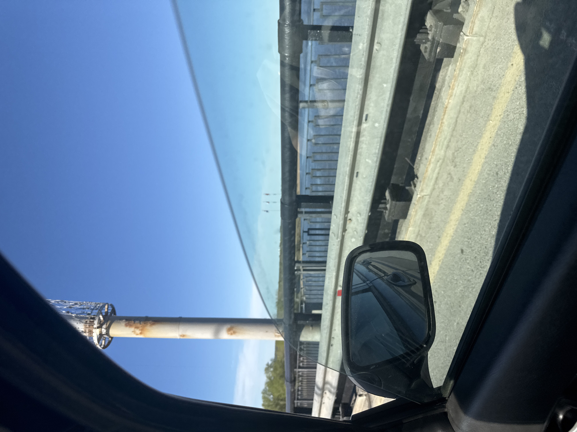 The image depicts a view from inside a vehicle, likely on a bridge, in daylight. The side mirror of the vehicle is visible, reflecting a section of the bridge. Outside the window, the sky is visible, along with a metal structure, possibly a lamppost or part of the bridge's infrastructure. The bridge's railing and concrete surface are also visible. The location is likely Perm, Russia, though this cannot be confirmed visually.