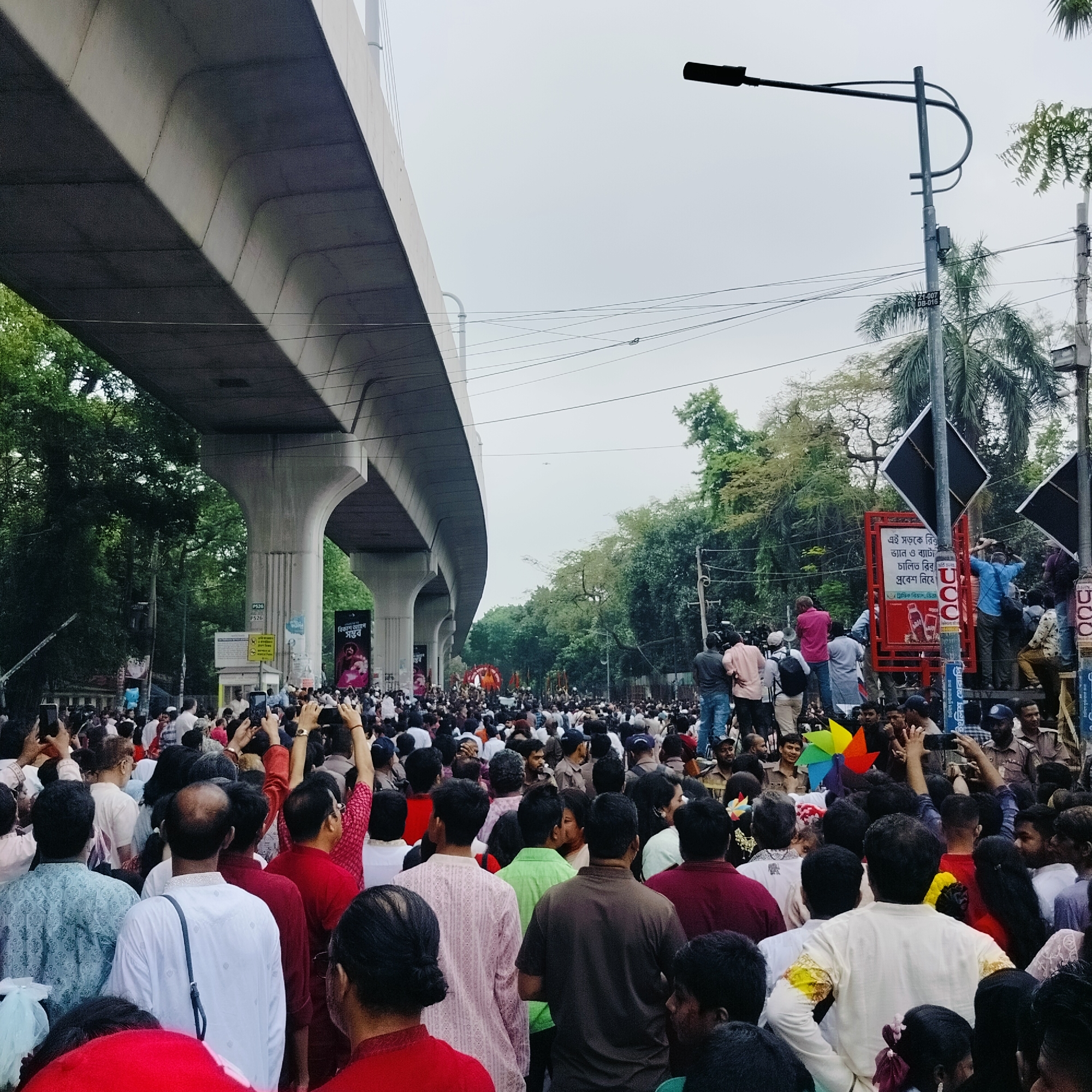 The image shows a large crowd of people gathered on a street in Dhaka, Bangladesh. An elevated road structure is visible above the crowd, supported by concrete pillars. A street lamp stands on the right. People are taking pictures with their phones. There are signs on the right, including one with "বিকাশ আগ্রপ সম্ভব" (Bikash Agrop possible). Other signs have traffic restrictions.