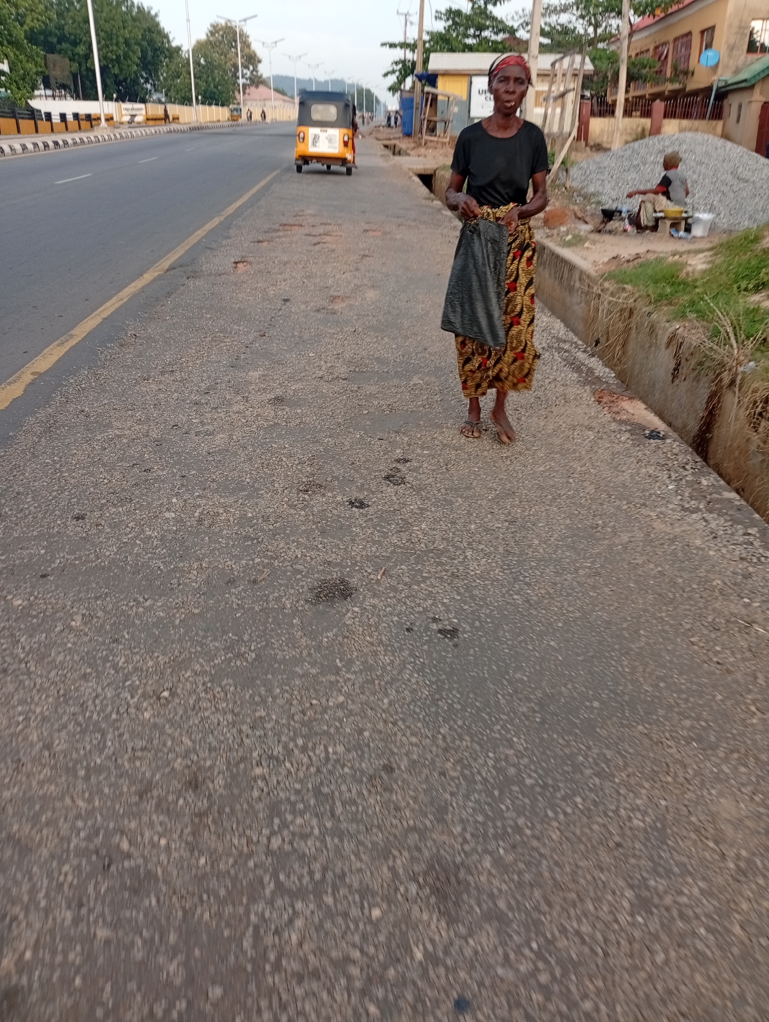 An elderly woman walks on the side of a road in Zango-ndaji, Nigeria. She is wearing a black shirt, a patterned skirt, and sandals. She is carrying a dark-colored bag. To her right is a drainage ditch and a patch of grass. Further away, behind her, is a person sitting on a stool, tending to a cooking station next to a pile of gravel. A yellow tuk-tuk drives down the adjacent road. The road has a yellow line dividing lanes. Beyond the road, buildings and trees are visible. The sky is overcast.