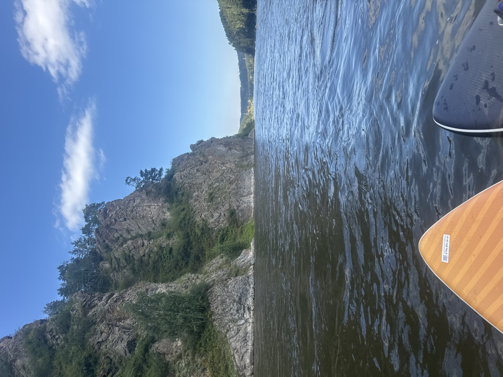 This is a first-person perspective shot from a paddleboard on a river in Ust'-Mana, Russia. In the foreground, the orange and striped tail of a paddleboard is visible on the left, and the dark blue tail of another paddleboard is visible on the right. The river is wide and dark, with ripples on its surface. On the left bank of the river, a steep, rocky cliff face rises, covered in sparse green vegetation and trees. The cliff is illuminated by bright sunlight, creating stark shadows. The sky above is a clear blue with a few scattered white clouds. In the distance, the river curves to the right, with tree-covered hills on both banks. A few structures and a bridge are visible in the far distance. The overall impression is of a serene natural landscape under a bright, sunny sky.