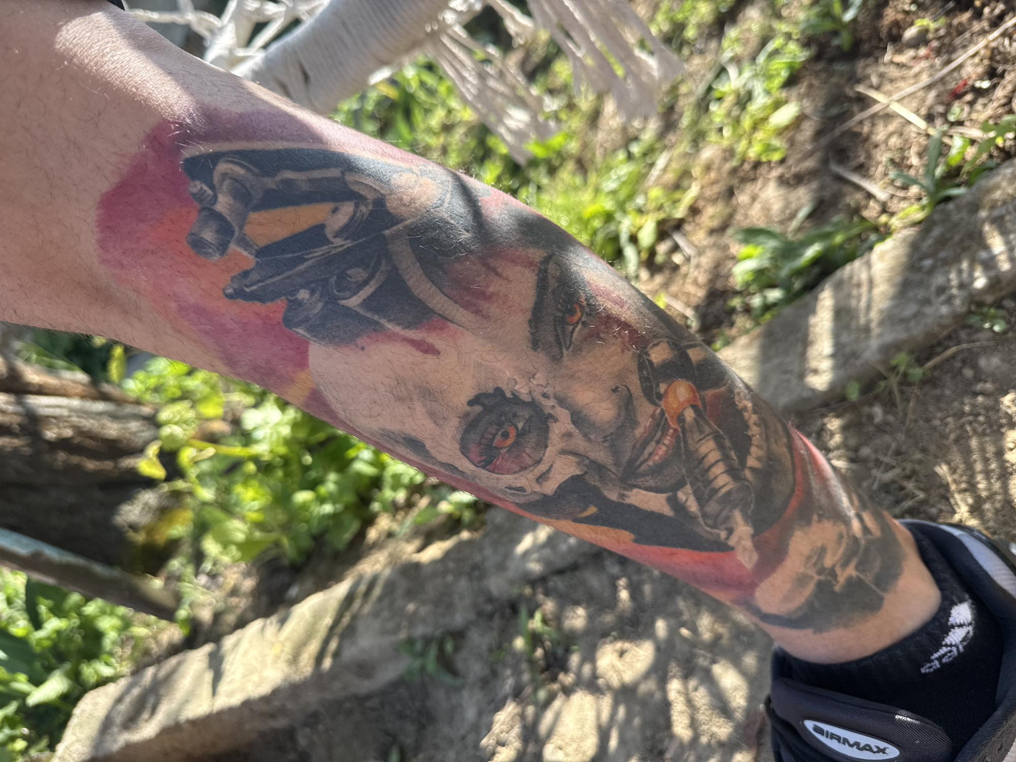 A human leg is shown in the foreground, featuring a complex tattoo with elements of machinery and facial features. The tattoo has a color palette of black, gray, and reds. The leg is positioned in front of an outdoor environment with green foliage and a section of a light-colored stone or concrete structure. At the bottom edge of the photo, a black sock and part of a shoe can be seen. The image appears to be taken outdoors in daylight. The location is Strazhitsa, Bulgaria.