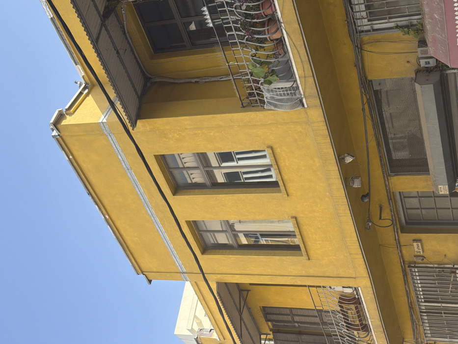 A multi-story building, painted bright yellow with areas of visible weathering and discoloration, is depicted under a clear blue sky. The upper facade features two levels. On the top visible floor, two large rectangular windows with dark frames reflect light, revealing white interior window components. A horizontal silver-colored trim band runs along the building's edge below the roofline and directly beneath these windows.
The floor below presents two balconies. The left balcony features a white ornate metal railing and closed brown louvered shutters, positioned beneath a corrugated metal awning. The right balcony also has a white ornate metal railing, below which a white bird deterrent spike strip is visible. This balcony contains multiple potted plants with red and green foliage, and a white, curved mesh object, possibly a cage or cover. A corrugated metal awning extends over this balcony as well.
Black electrical cables traverse the facade horizontally below the balconies, connecting to several small grey utility boxes. Two small birds are perched on these wires or fixtures. A yellow rectangular sign, displaying "30 phon" in black text, is affixed to the wall below the left balcony. The partially visible ground floor includes windows with metal grilles and a grey roll-down commercial shutter. A dark red awning is visible on the far bottom right. A lighter-colored building is partially visible behind the yellow structure on the left. The scene is located in Tel-Aviv, Israel.