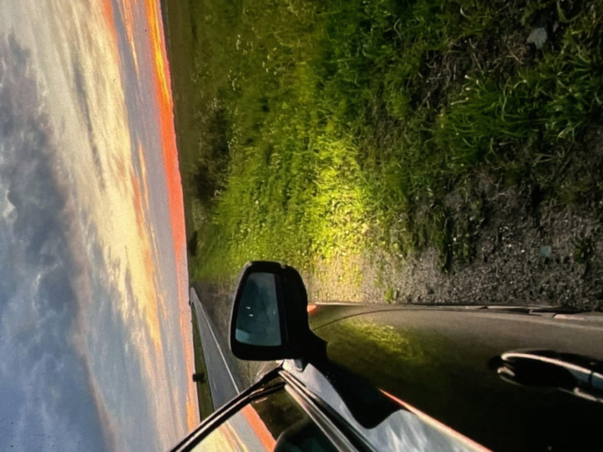 The image depicts a roadside view at dusk, with a car parked on the right side of a paved road. The car's side mirror and a portion of its dark body are visible in the foreground, reflecting the surrounding environment. The road stretches into the distance, curving slightly, with a green, grassy field on the right side of the road. The field is illuminated by a bright, yellowish light originating from the car, highlighting the texture of the grass and small white flowers. The sky above is a dramatic display of sunset colors, with bands of orange, pink, and yellow mingling with darker gray clouds. A single, isolated tree can be seen in the middle distance on the left side of the road. The overall scene conveys a sense of a rural or semi-rural landscape. This image was captured in the vicinity of Harmanli, Bulgaria.