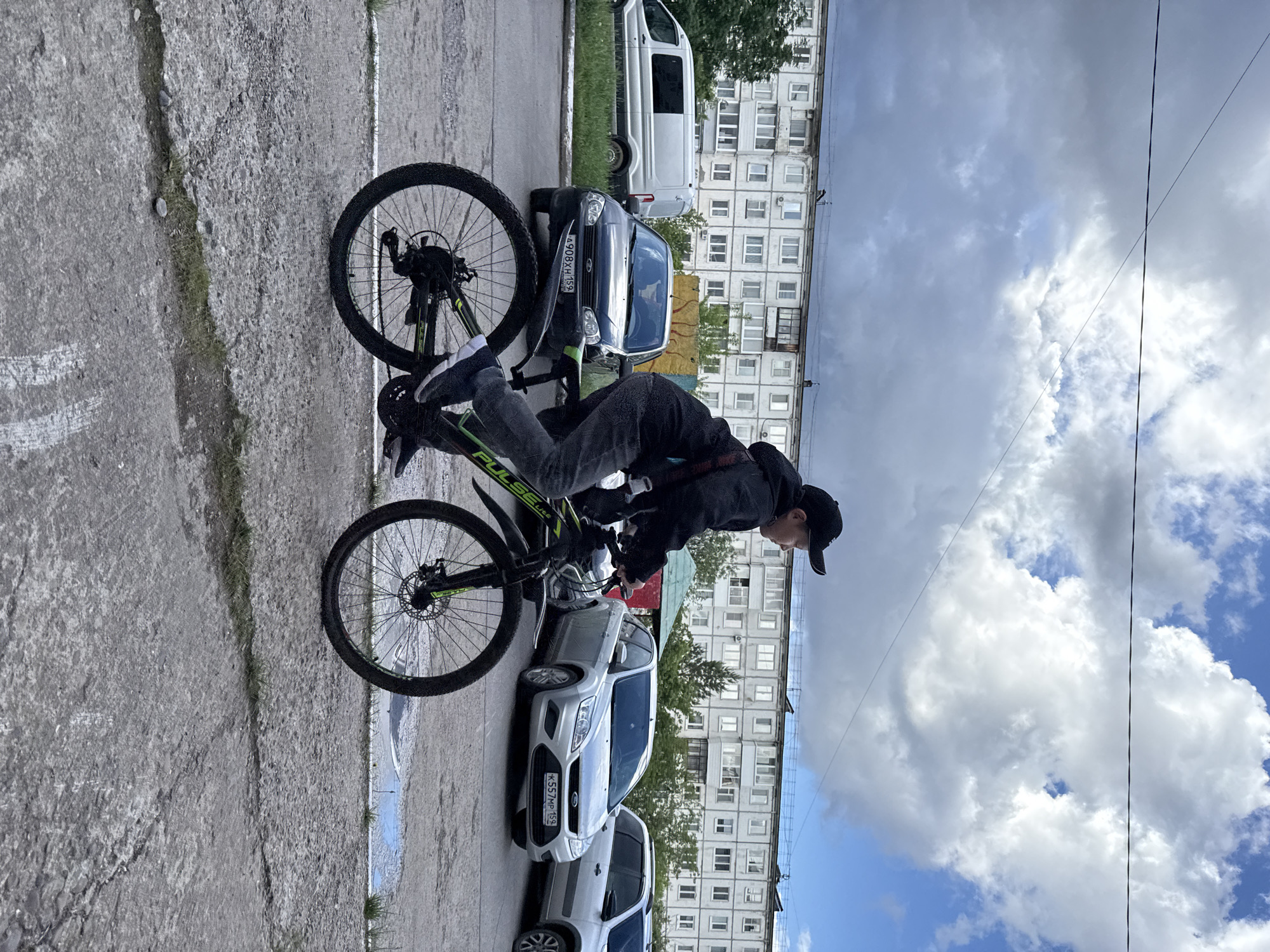 The image depicts a young person on a bicycle on a partially cloudy day in Chaikovsky, Russia. The individual, wearing a dark cap, a dark jacket, and jeans, is positioned on a green and black "PULSE Lite" bicycle, leaning forward over the handlebars as if in motion or preparing to ride. A "NIKE" logo is partially visible on an item of clothing or a bag worn by the person.
The scene is set on a concrete paved surface, likely a sidewalk or a side street. Along the adjacent street, several cars are parked. A dark blue car bears the Russian license plate A908XH159, and a silver Ford Focus has the license plate K557MP152. A white van and another silver car are also visible further down the street.
In the background, multi-story residential apartment buildings, typical of Soviet-era architecture, line the street. Patches of green grass grow along the curb and between the pavement and the buildings. The sky above is distinctly cloudy with large white clouds, but also shows clear patches of blue, indicating it is daytime. The concrete ground exhibits cracks and some small weeds.