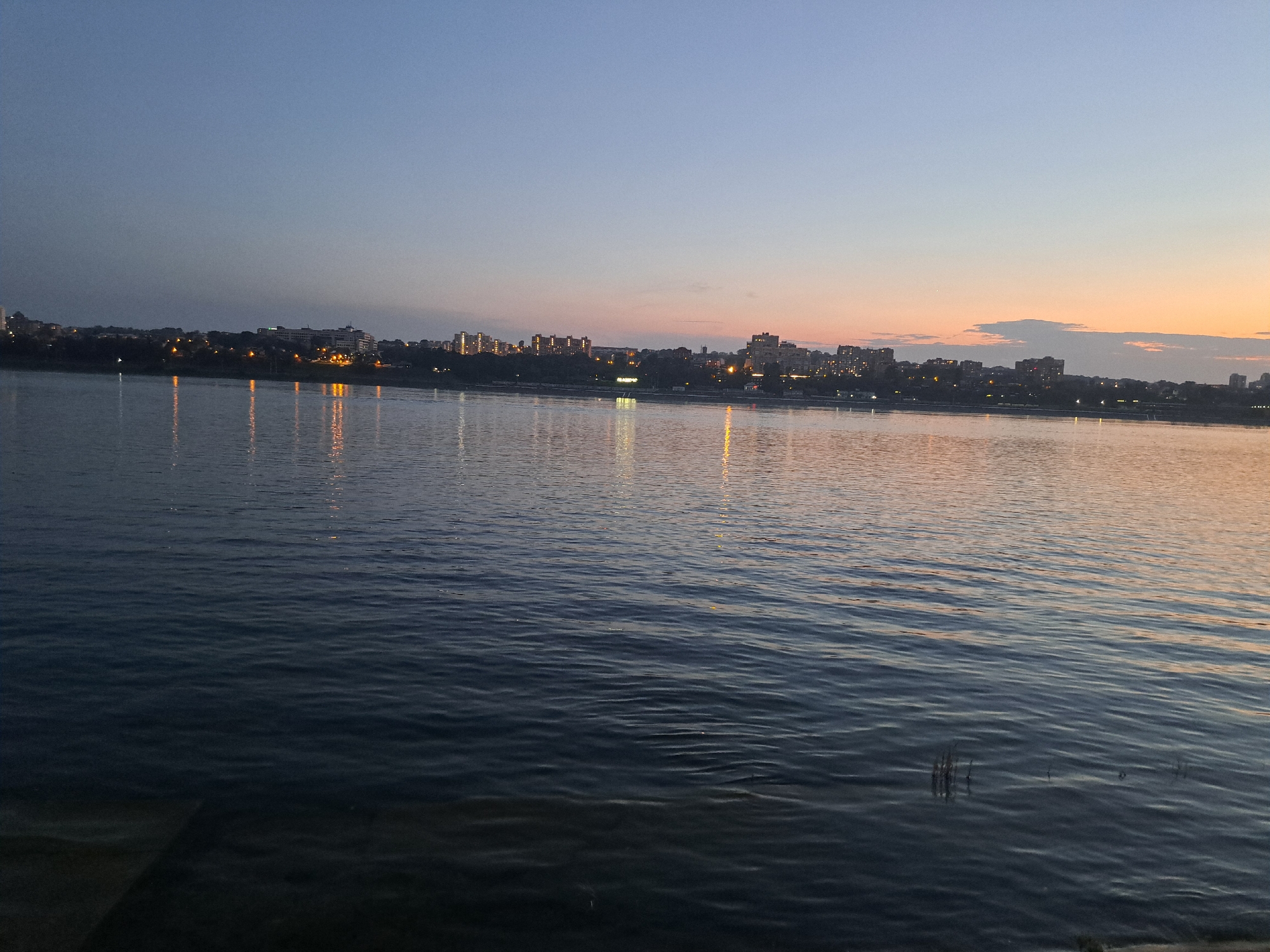This image captures a serene twilight view of the Angara River in Irkutsk, Russia. The water's surface is calm, reflecting the faint lights of the city skyline that stretches across the horizon. The sky displays a gradient of colors, transitioning from a deep blue at the top to softer hues of pink and orange near the horizon, characteristic of a sunset or sunrise. The city lights are visible as small, bright dots scattered along the distant shore, interspersed with the darker silhouettes of buildings and trees. The foreground shows the dark, rippling water, with some subtle textures and shadows indicating depth or submerged objects. The overall atmosphere is peaceful and tranquil, evoking a sense of calm as day transitions into night.