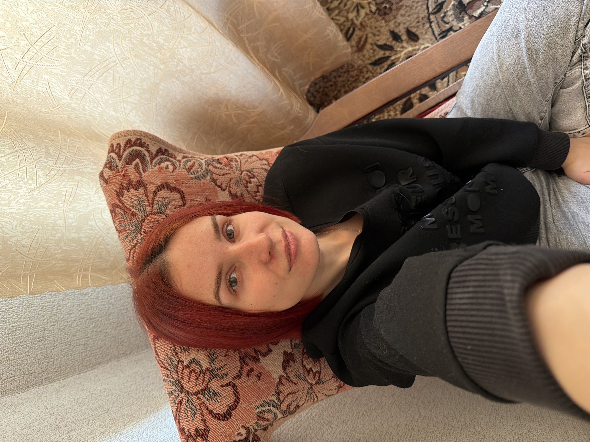 A fair-skinned woman with red hair is reclining on an ornately patterned chair in an indoor setting. She is wearing a black sweatshirt and gray jeans. The chair is positioned next to a couch and a decorative rug. The location is within an interior space, possibly in Chaikovsky, Russia.