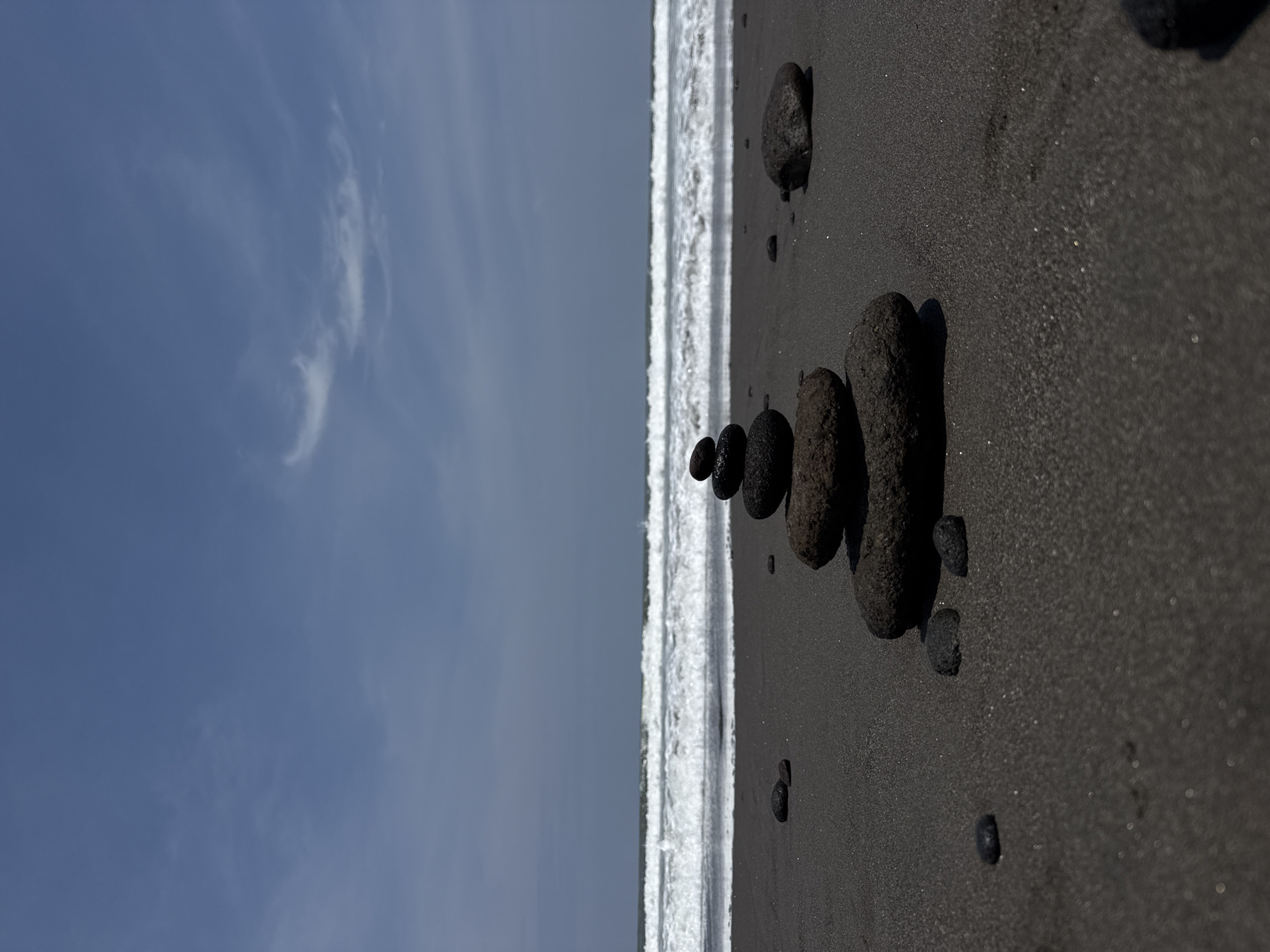 The scene depicts a black sand beach in Sudimara, Indonesia, under a clear to partly cloudy blue sky. In the foreground, a prominent cairn composed of five dark, smooth, igneous rocks is stacked centrally on the dark, fine-grained sand. Scattered smaller, similarly dark rocks are visible on the sand around the cairn. The midground features the ocean with multiple parallel lines of breaking white-capped waves rolling towards the shore. The background consists of a light blue sky with wispy white cloud formations, including a distinct, elongated, curved cloud centrally positioned. No individuals or man-made structures are visible. The environment appears natural and undisturbed by human activity, with natural light indicating daytime conditions.
