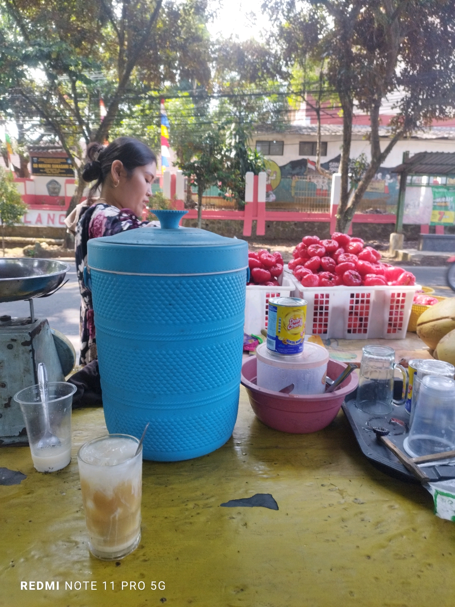 The image depicts an outdoor scene in Saruni, Indonesia, likely a roadside stall. A woman, with dark hair pulled back and wearing a floral-patterned top, is partially visible, appearing to be engaged in an activity related to the stall. In the foreground, a large blue insulated cooler stands prominently on a yellow table. Several glasses are present; one contains a milky beverage with ice and a spoon, and another is partially filled. Also on the table are plastic cups, a metal bowl, and a can of condensed milk. Behind the cooler, two white baskets overflow with bright red fruit, possibly rose apples. Coconuts are also visible. The background shows trees, buildings with red and white accents, and a fence, suggesting an urban or semi-urban environment. The sunlight indicates it is daytime.