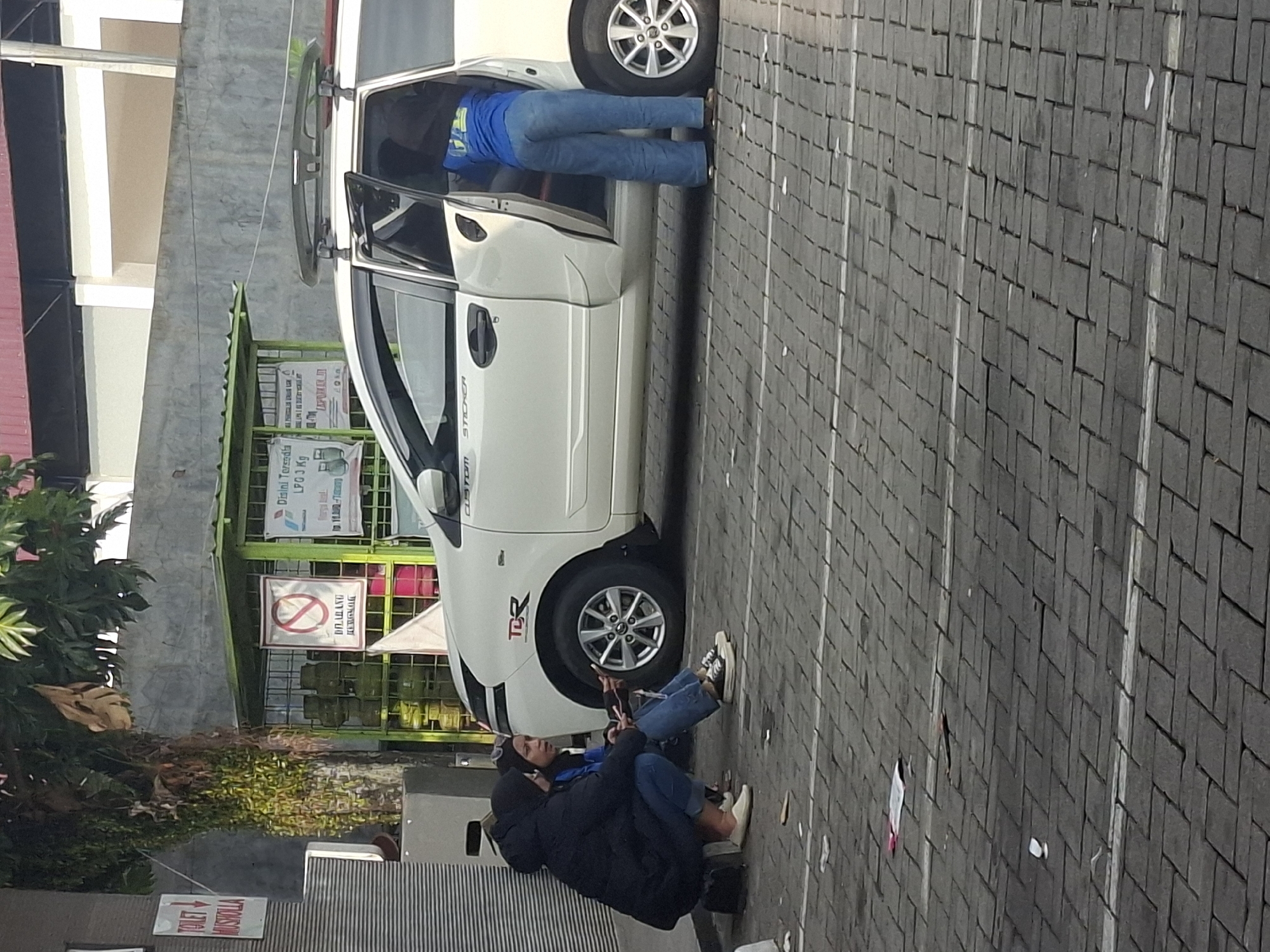 Two individuals, appearing to be women, are seated on the ground next to a white minivan in Cikarang, Indonesia. The individual on the left is wearing a dark hooded jacket and blue jeans, while the person on the right is wearing a blue jacket and blue jeans. They are both looking towards the left. The minivan's driver-side door is open, and a third person, wearing a blue shirt and blue jeans, is standing inside the vehicle, partially visible from behind. A rack is mounted on the roof of the minivan. In the background, a green metal cage holds several propane gas cylinders. A sign with a red circle and a diagonal line is attached to the cage, indicating a prohibition. Adjacent to the gas cylinders is another sign with text in Indonesian. The ground is paved with interlocking bricks. Debris, including paper and a white cup, is scattered on the ground.