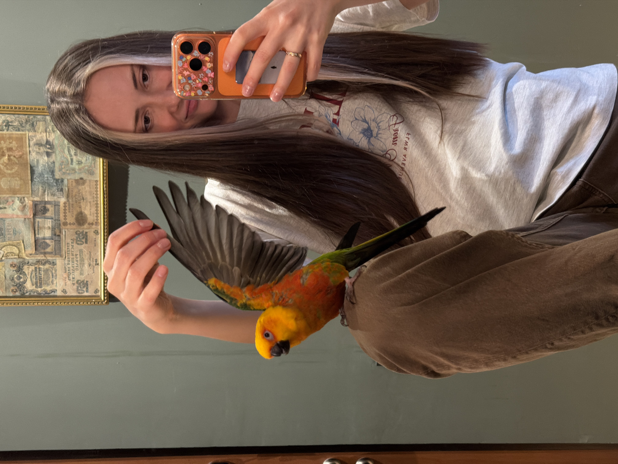 A young woman is reflected in a mirror as she takes a selfie with her phone. She is wearing a light gray t-shirt with a floral design and brown pants. She has long brown hair with a blonde streak. A brightly colored Sun Conure parrot is perched on her leg, with its wing extended toward the woman's hand. A framed piece of artwork hangs on the wall behind her. The location appears to be indoors, possibly a home. There is no information about Khimki, Russia in the image.