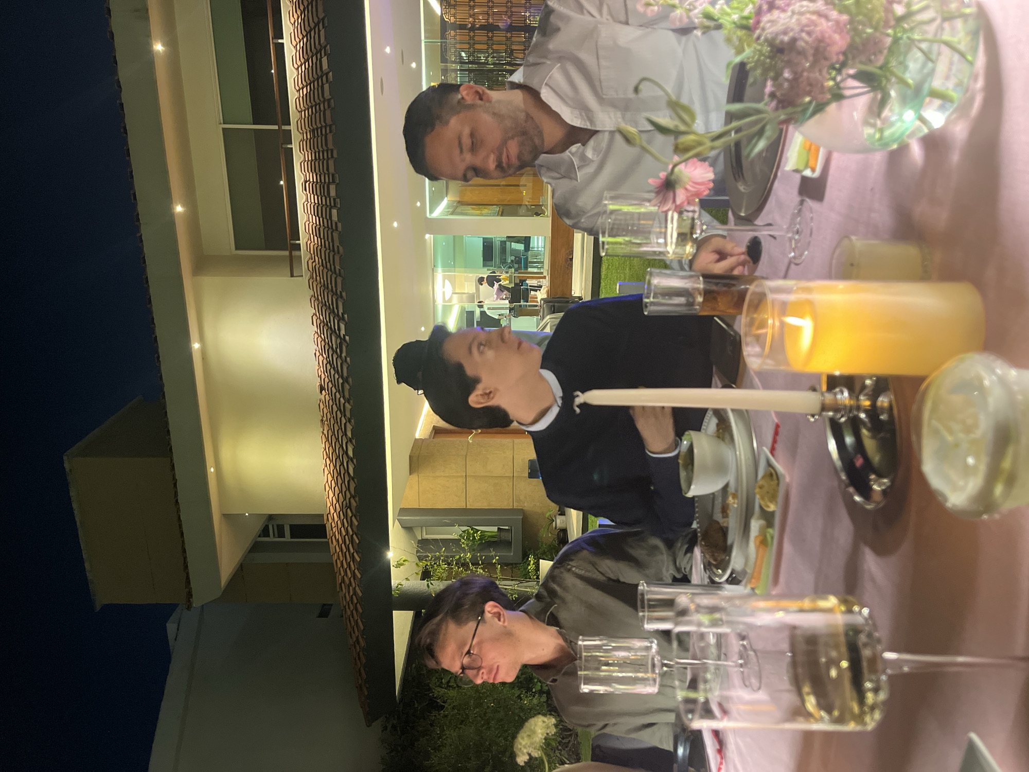 The image depicts three individuals seated at a dining table. A man in a white shirt is positioned at the top of the frame. A person in a dark jacket sits in the center. Another individual wearing glasses and a gray shirt is at the bottom. The table is set with candles, flowers in a vase, plates, and an arrangement of wine glasses. The location appears to be outdoors, adjacent to a building. Based on the architecture of the building, it is likely that the picture was taken in San Bernardino Tlaxcalancingo, Mexico.