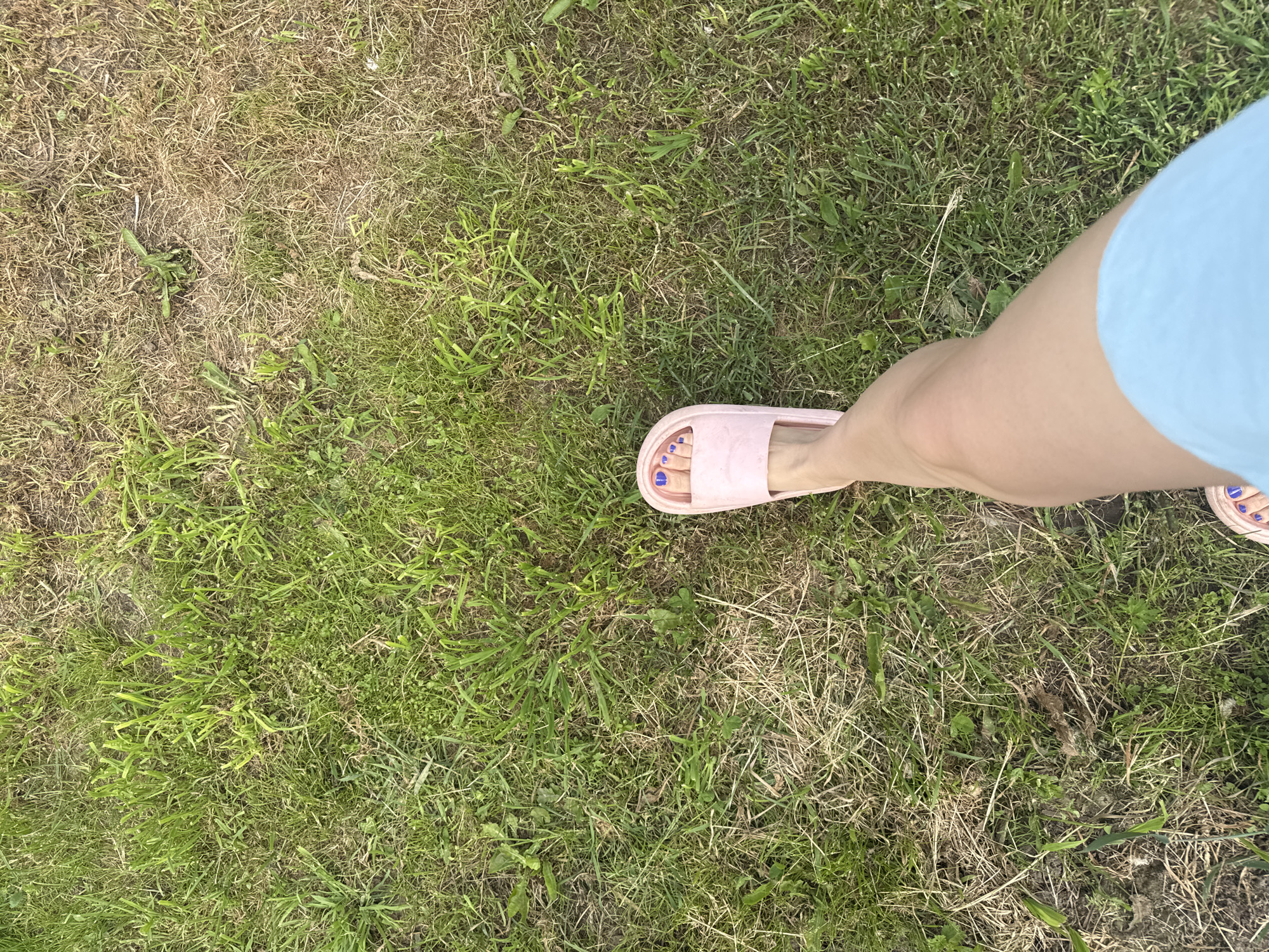 The image shows a close-up, high-angle view of a person's left leg and foot standing on a grassy area. The person is wearing a light blue garment, possibly shorts or a skirt, and a pink slide sandal on their left foot. The sandal reveals toes painted with blue nail polish. The ground is covered in a mix of green and dry grass, with some patches of bare earth. The overall scene suggests an outdoor setting. The location is described as Луптюг, Russia.