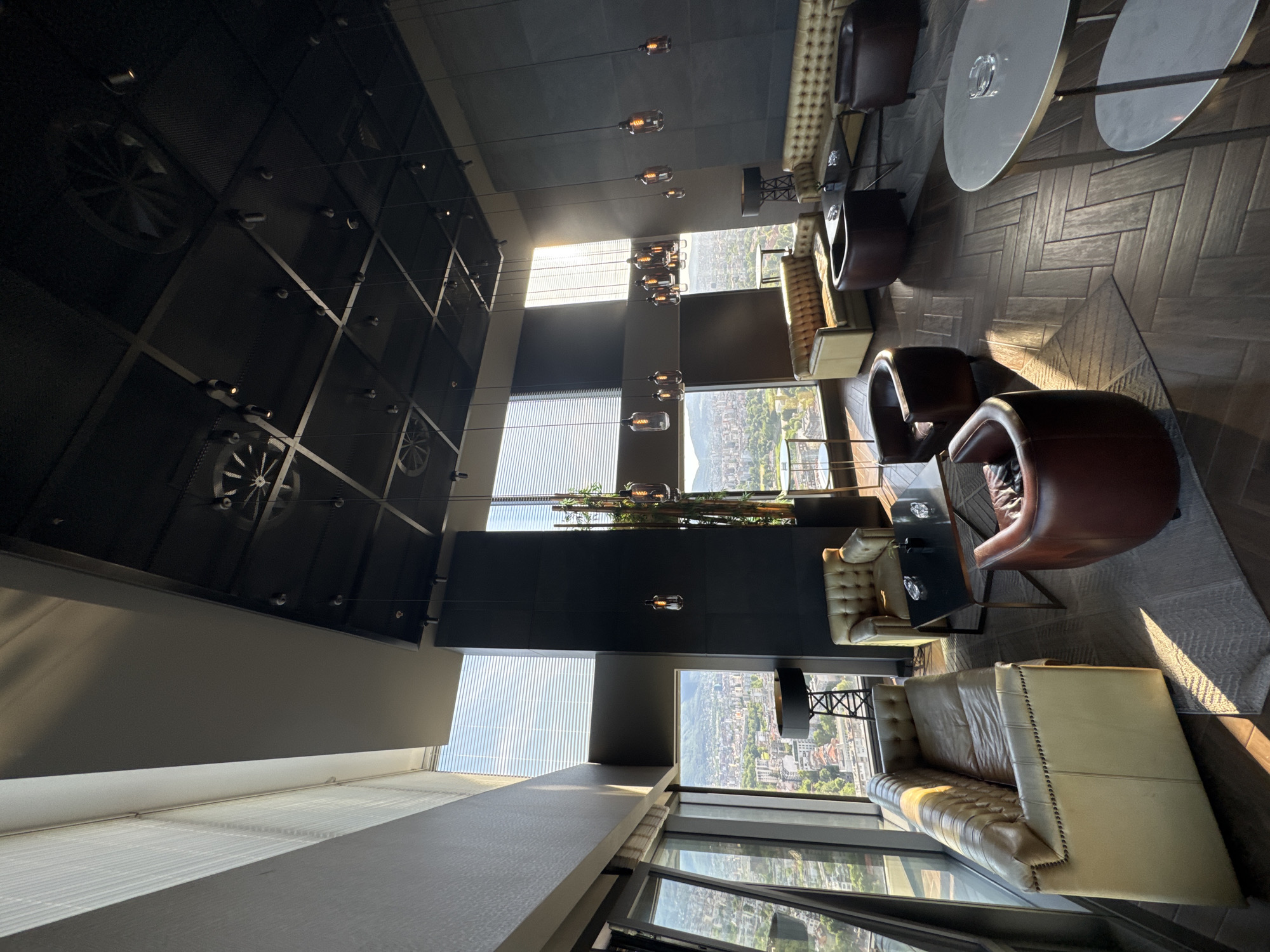 The image depicts a high-rise interior, likely a bar or lounge, with large windows overlooking a city. The dominant color scheme is neutral with browns, grays, and whites. Furnishings include leather armchairs and sofas, and tables with reflective surfaces, set on a herringbone patterned floor. The ceiling features decorative mechanical-themed panels with integrated lighting. Vertical blinds partially cover the windows, obscuring the view of the cityscape, but the location is Sofia, Bulgaria. No people are visible in the frame.