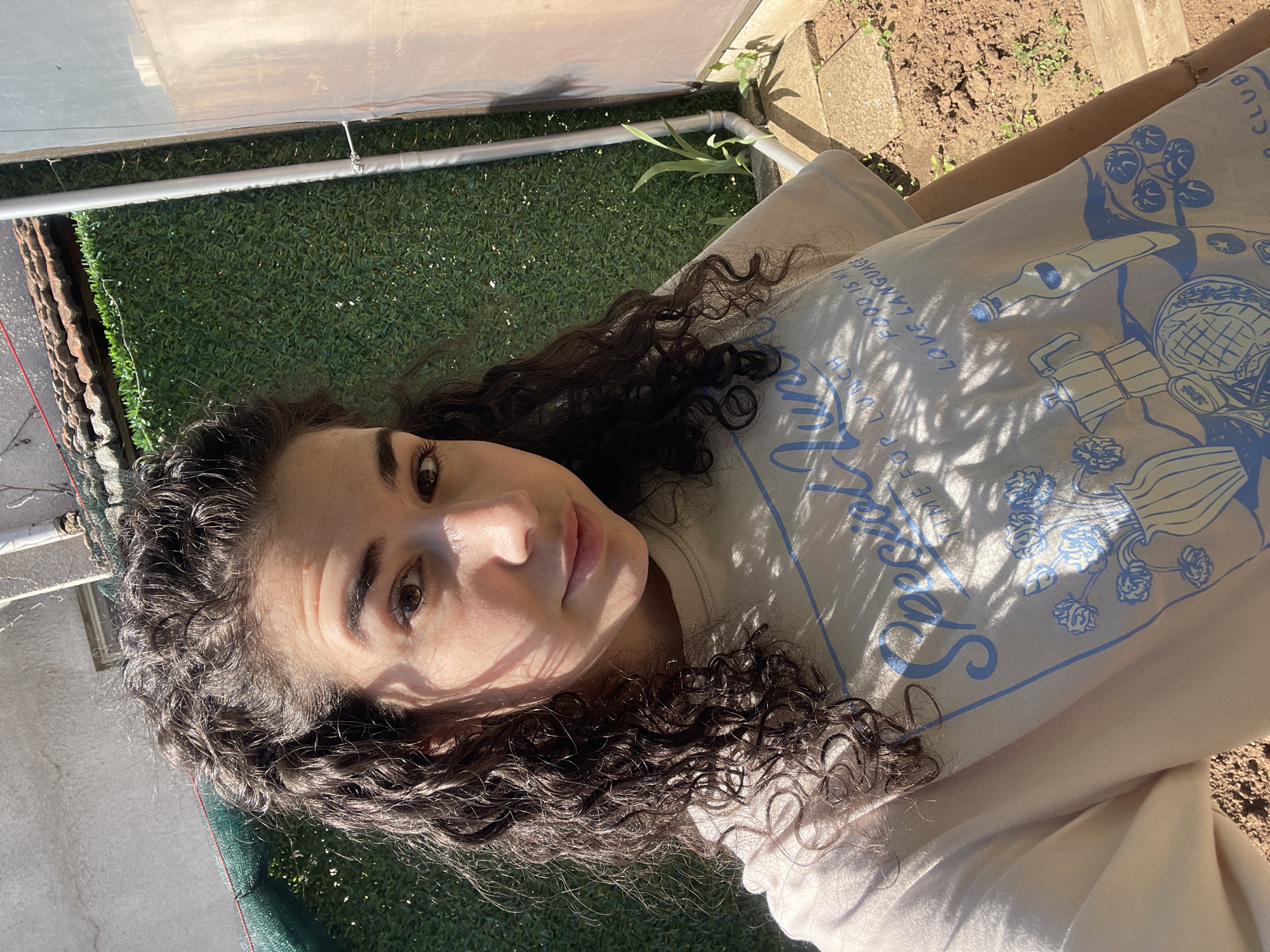 A woman with curly dark hair lies on a beige surface in an outdoor setting. She is wearing a light-colored top with blue graphics that feature images of kitchen items and the words "Special time." Sunlight filters through, casting shadows across her face and clothing. The background features a green wall or artificial turf, and a covered structure with metal supports. Patches of dirt and small plants are visible near a concrete border, suggesting a garden or patio area. Without additional information or location data, it is impossible to determine if the photograph was taken in Sofia, Bulgaria.