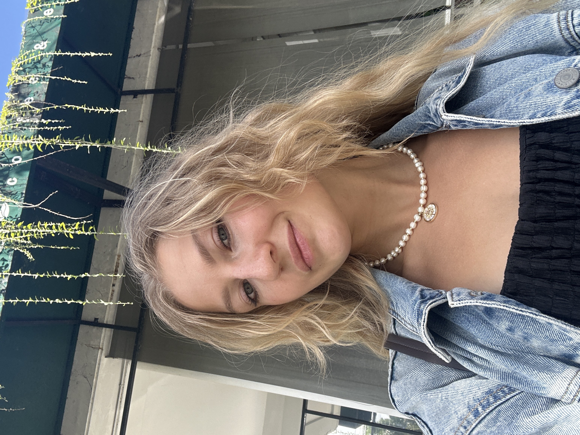 A young woman poses for a selfie. She has blonde, wavy hair and is wearing a pearl necklace with a gold pendant and a light-wash denim jacket over a black top. The background includes building exterior and vegetation. The location cannot be determined from the image. The city of Kuta Selatan, Indonesia cannot be confirmed.