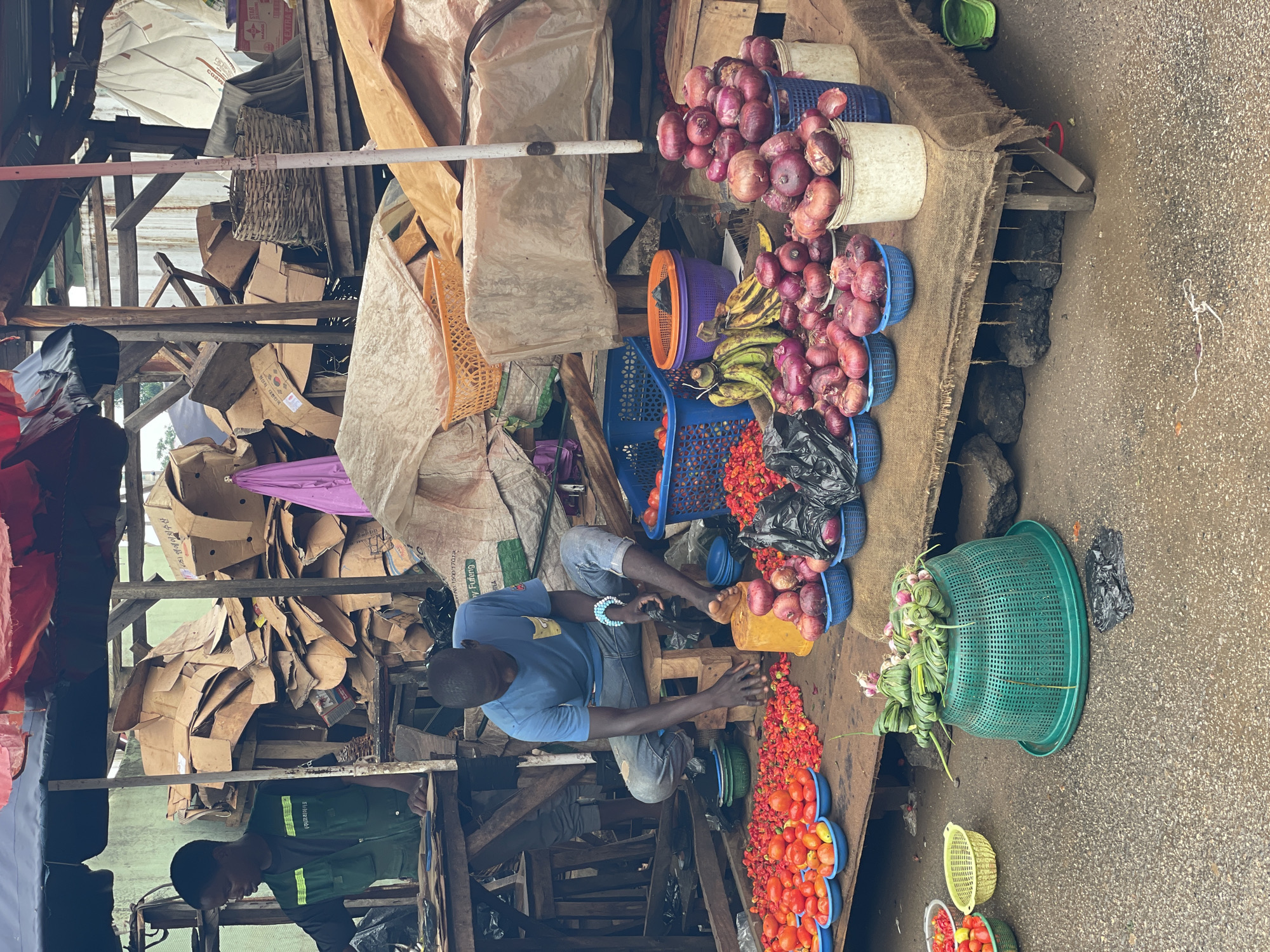 The image depicts a street market scene in Akwanga, Nigeria. In the foreground, a man is seated on a wooden stool, sorting through red chilies. He is wearing a blue t-shirt and blue jeans. In front of him, various produce is arranged in baskets and on the ground, including tomatoes, red chilies, onions, and bananas. A large green basket filled with bundled greens sits on the ground to the left. Behind the man, a second person, wearing a green vest over a dark shirt, is visible behind a stall. The background is cluttered with cardboard boxes and tarpaulin coverings, suggesting the market is partially covered. The ground appears to be asphalt, and there are some rocks and debris scattered around. The overall impression is of a busy, open-air market.