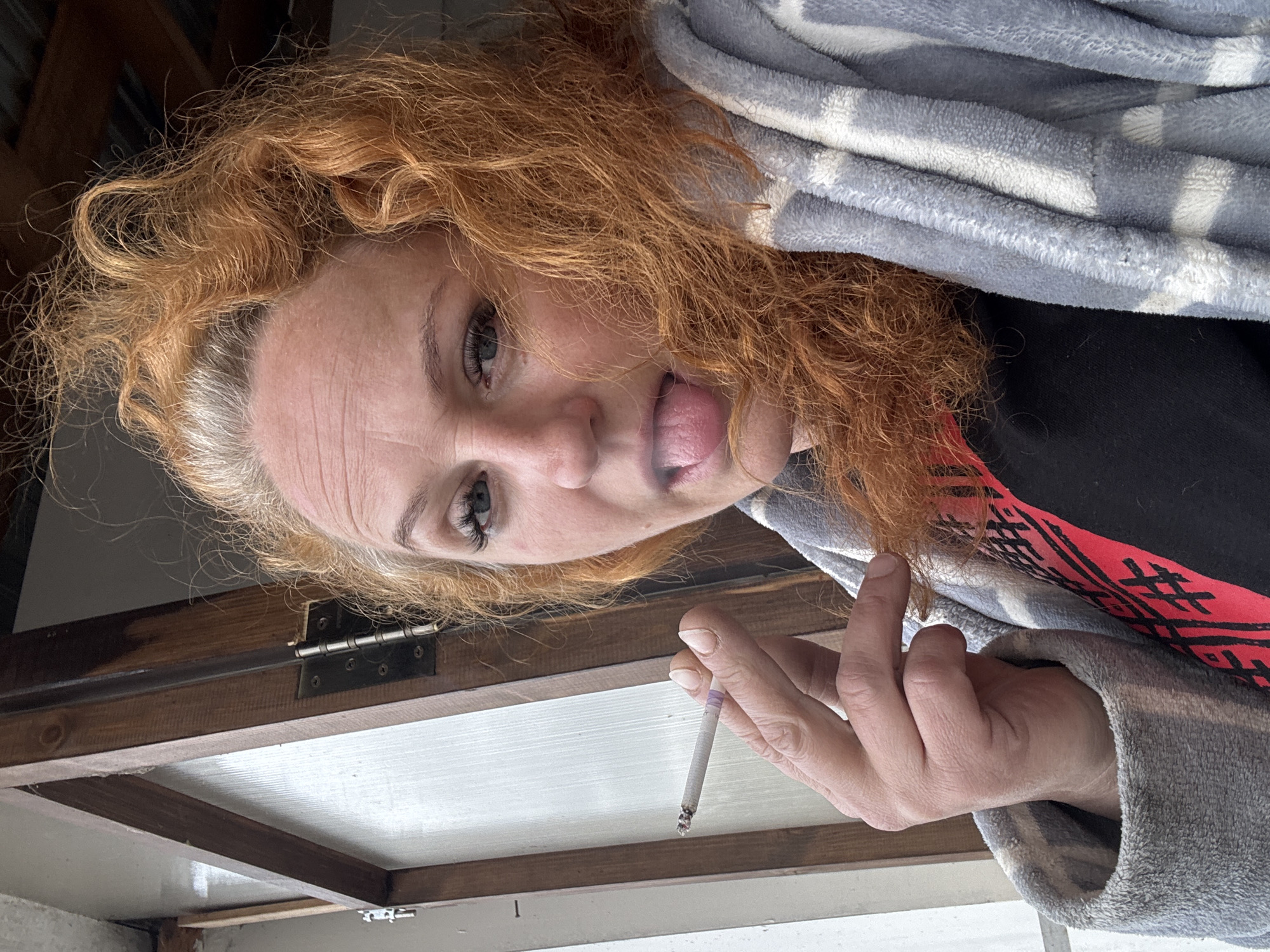 The image is rotated approximately 90 degrees clockwise, depicting a woman's upper body and head captured from a low angle, looking upwards into the camera. She has voluminous, wavy red hair with visible graying strands near her temples. Her expression is playful, as she sticks out her tongue. Notable facial features include light-colored eyes and visible wrinkles around her eyes and on her forehead.
The woman is wearing a plush, grey and white patterned robe or blanket. Beneath it, a black garment is visible, which appears to have a red and black patterned design on what looks like a sleeve or an underlayer. In her right hand, she holds a lit cigarette with a distinctive purple filter, and her fingernails are neatly manicured.
The background suggests an indoor setting, featuring a substantial wooden structure with visible beams and a metal hinge positioned directly behind and above her. This could be part of a window frame, a ceiling element, or furniture. Below this wooden section, there is a lighter, possibly white or light grey, surface. The ambient lighting indicates that the photograph was taken during the daytime. There is no visible text in the image. The overall mood conveyed is informal and candid.