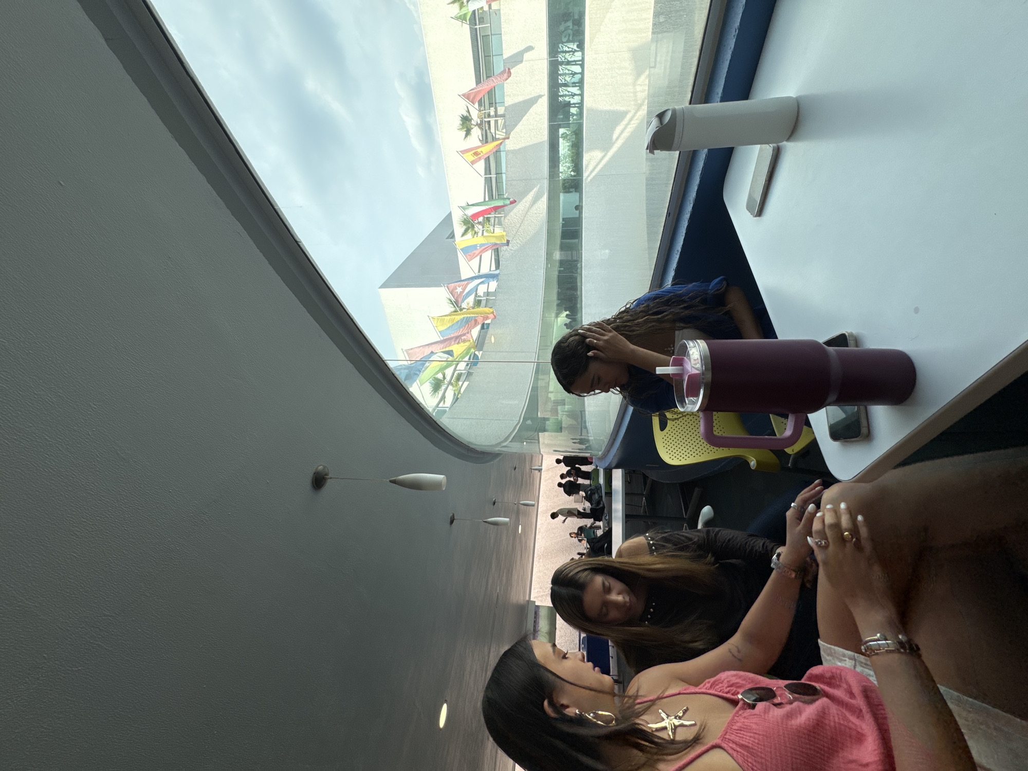 Three young women are seated at a white table in a well-lit indoor space. One woman wears a pink top with a starfish necklace and has sunglasses resting on her chest. Another wears a black top with studded detailing. The third woman wears a blue top. All three women appear to be engaged in conversation. A large maroon-colored tumbler and a white water bottle are placed on the table. Outside the large window are multiple flags and the architectural features of the Complejo Cultural Universitario de la BUAP in Heroica Puebla de Zaragoza, Mexico. Other people are seen walking in the background through the window.