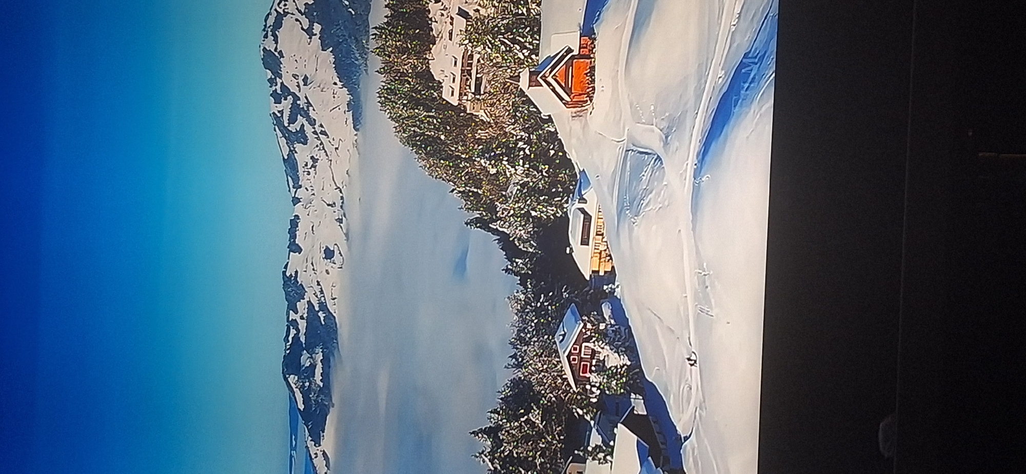 The image shows a serene winter landscape with snow-covered mountains and hills. In the foreground, several houses are nestled amongst snow-laden pine trees. The houses appear to be made of wood, with gabled roofs, and some have warm lighting visible through the windows. The terrain is sloped, with ski tracks visible in the snow, suggesting recreational activity. A layer of fog or clouds covers the lower parts of the mountains in the background, creating a sense of depth. The sky is a clear, bright blue. There is no indication of the city of Bila Tserkva, Ukraine, in this image.
