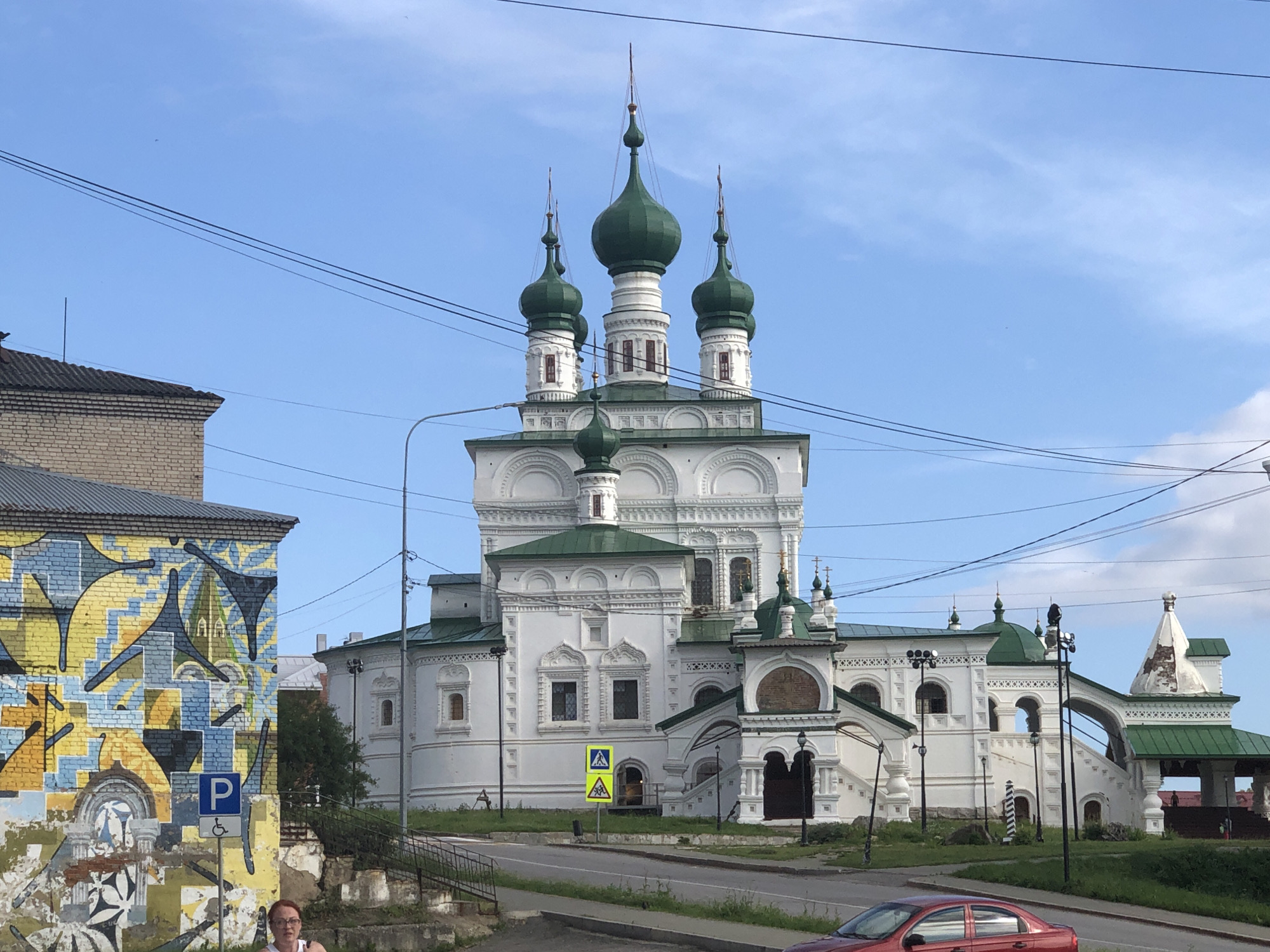 This media file captures a view of a white church with multiple green domes and spires against a clear blue sky. The church is ornate with decorative white architectural elements. In the foreground, to the left, is a building with a vibrant mural on its brick wall, featuring abstract yellow, blue, and black shapes. A street runs in front of the church, with a few cars and some greenery.
A woman with red-rimmed glasses and a white top is partially visible in the lower left corner, looking towards the camera. To her right, a parking sign with a blue "P" and a symbol for disabled access is visible on a pole, mounted on the muraled building. Yellow pedestrian crossing signs are also present on the street in front of the church.
The scene appears to be set during the daytime, with bright sunlight casting shadows, suggesting a fair-weather day. The architecture of the church, with its distinct domes and intricate facade, along with the presence of Cyrillic-style street signs, strongly suggests a location in Russia, consistent with the provided context of Solikamsk. The overall atmosphere is peaceful and scenic. There is no visible text on the church itself, but street signs are present.