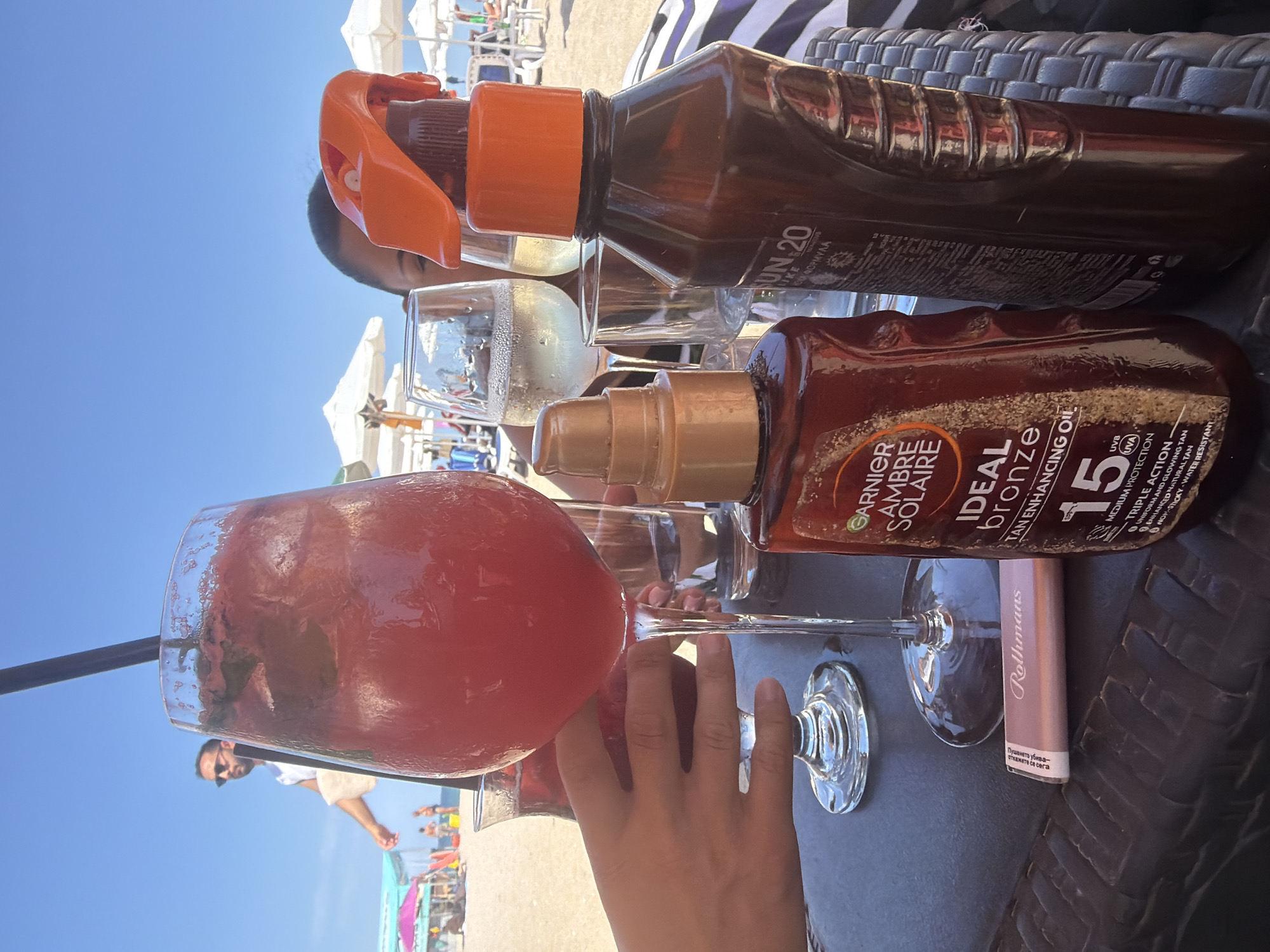 The image depicts a close-up scene on a beach in Sozopol, Bulgaria, focusing on a table at what appears to be a beach bar. In the foreground, two bottles of sun care products are prominently displayed: a "Garnier Ambre Solaire Ideal Bronze Tan Enhancing Oil SPF 15" and another bottle with "SUN.20" visible on its label, likely a sunscreen. A pink tube, possibly lipstick or a similar cosmetic, is placed on the table. A hand, belonging to a person out of frame, is reaching towards a large glass containing a red beverage with ice and mint. Behind these items, two glasses of white wine are visible. In the background, the beach is crowded with people relaxing under umbrellas, with the clear blue sky overhead. A man in sunglasses is walking on the beach in the distance. The overall setting suggests a leisurely day at the seaside.