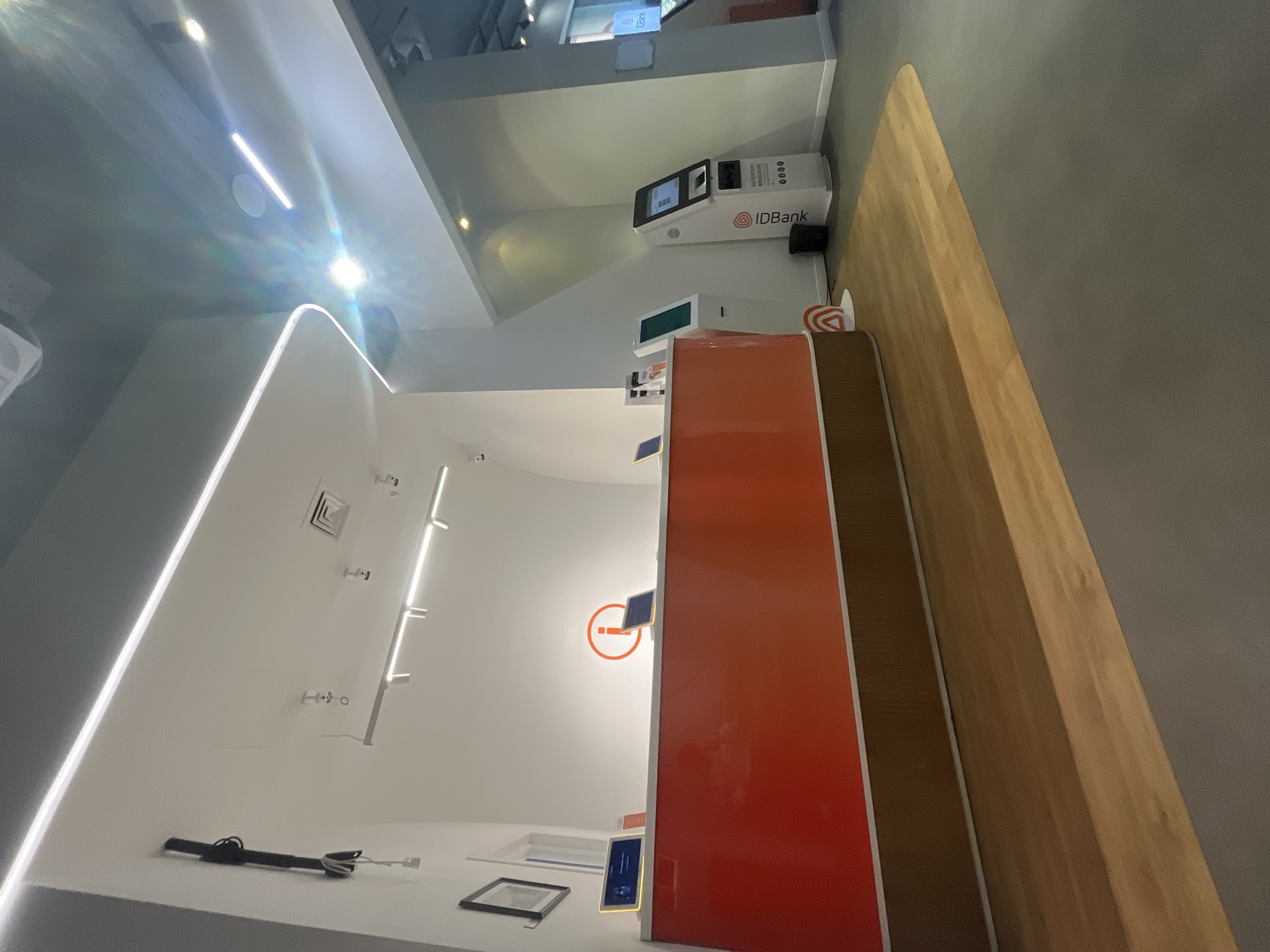 The image depicts the interior of an IDBank branch, likely located in Yerevan, Armenia, given the presence of IDBank branding on an ATM. The space features a modern design with a white interior, illuminated by linear LED lights. Visible elements include an orange partition, a wooden countertop, a Supercash ATM, wall-mounted informational displays, and security cameras. The scene is devoid of people.