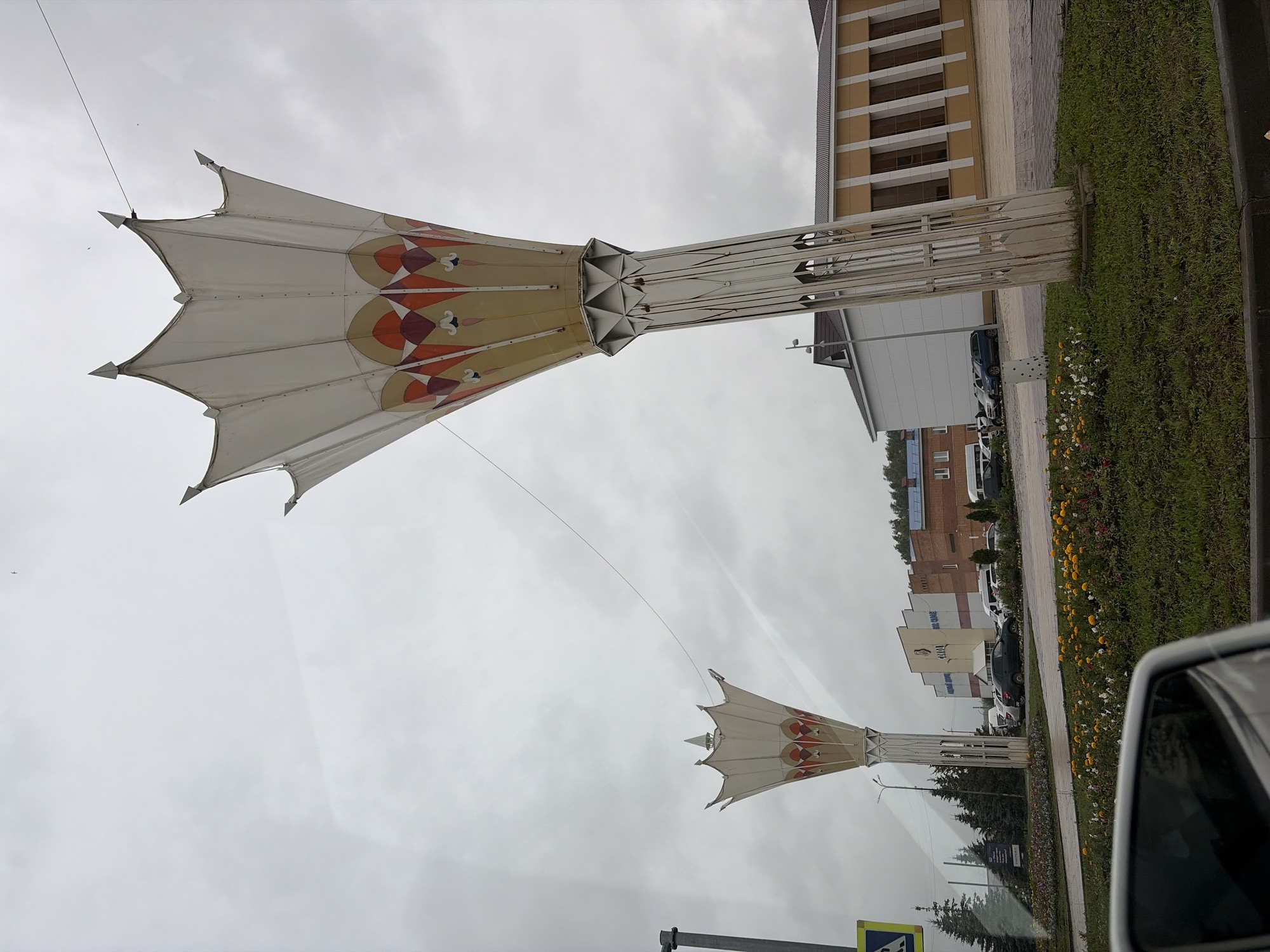 The image depicts an outdoor scene on an overcast day, likely in Aznakayevo, Russia. The primary subjects are two large, decorative umbrella-like structures mounted on tall, metal poles. These structures have a white fabric canopy with a repeating geometric pattern in shades of orange, red, and beige. The canopy is supported by a spiky, multi-pointed edge.
In the background, there are several buildings, including a light-brown building with a repeating pattern of windows and a white building. A dark-colored car is parked near the sidewalk. A strip of green grass with some scattered white and yellow flowers runs along the side of the sidewalk. The sky is a uniform gray, suggesting cloudy weather. A thin wire stretches across the sky from the top of the nearest umbrella structure. In the distance, a faint airplane can be seen in the sky.
The scene appears to be a public space, possibly a street or a plaza, given the presence of the sidewalk and the decorative structures. There are no people visible in the image. The overall mood is subdued, likely due to the cloudy weather. No discernible text is present in the image.